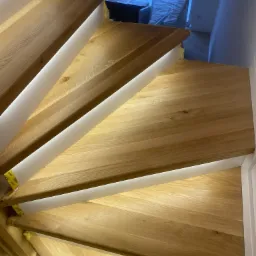 Bieg drewnianych schodów z podświetleniem LED, widok z góry, ukazujący naturalny rysunek drewna i precyzyjne wykonanie, z elementami wykończenia w bieli.