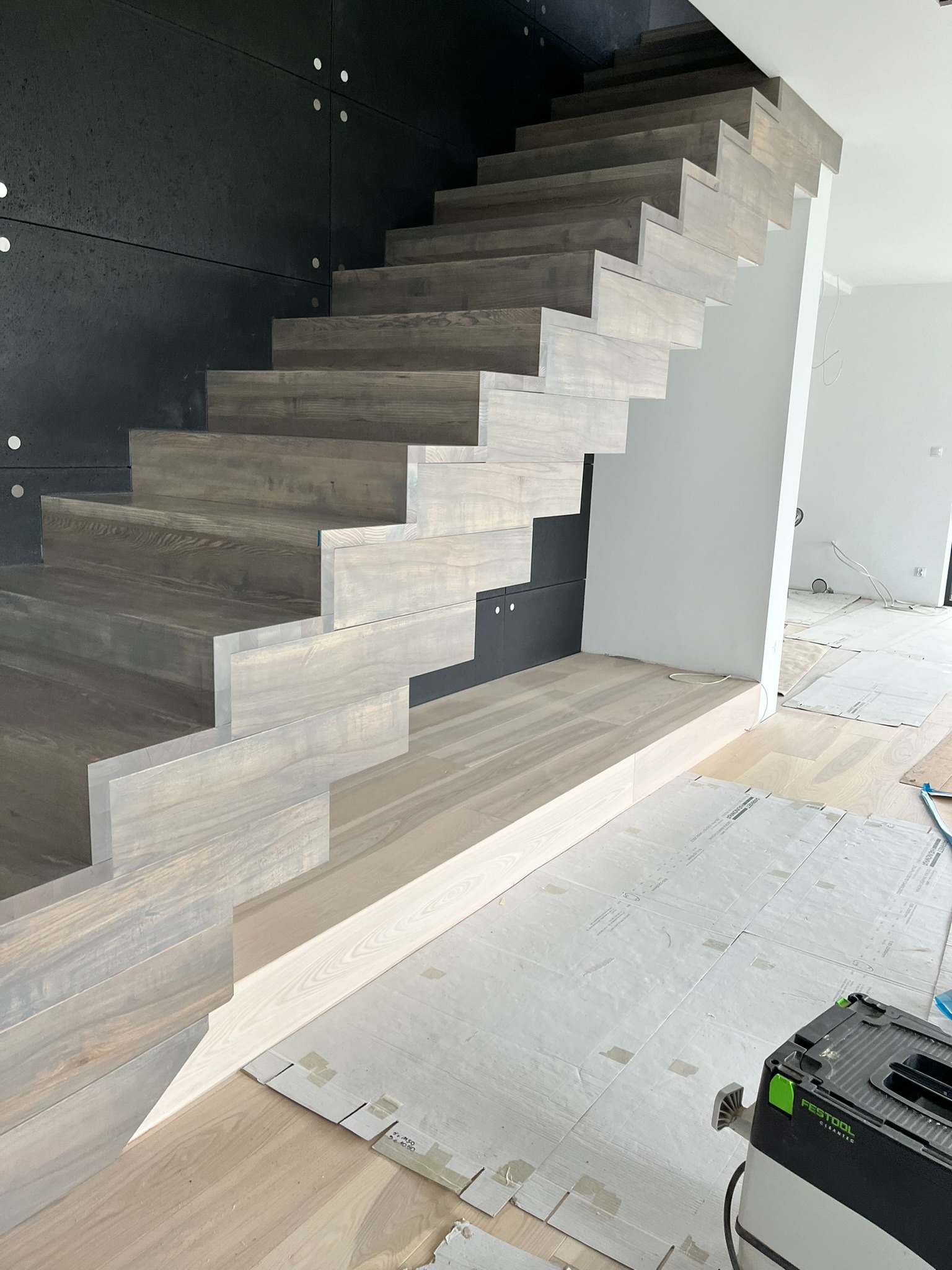 Nowoczesne, minimalistyczne schody drewniane w jasnej tonacji, z ciemnoszarą ścianą w tle. Widoczny odkurzacz przemysłowy i zabezpieczona podłoga.