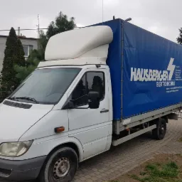 Biały samochód dostawczy Mercedes-Benz Sprinter z plandeką reklamową firmy HAUSBERGER ELEKTROTECHNIK, zaparkowany na kostce brukowej, widok z boku.