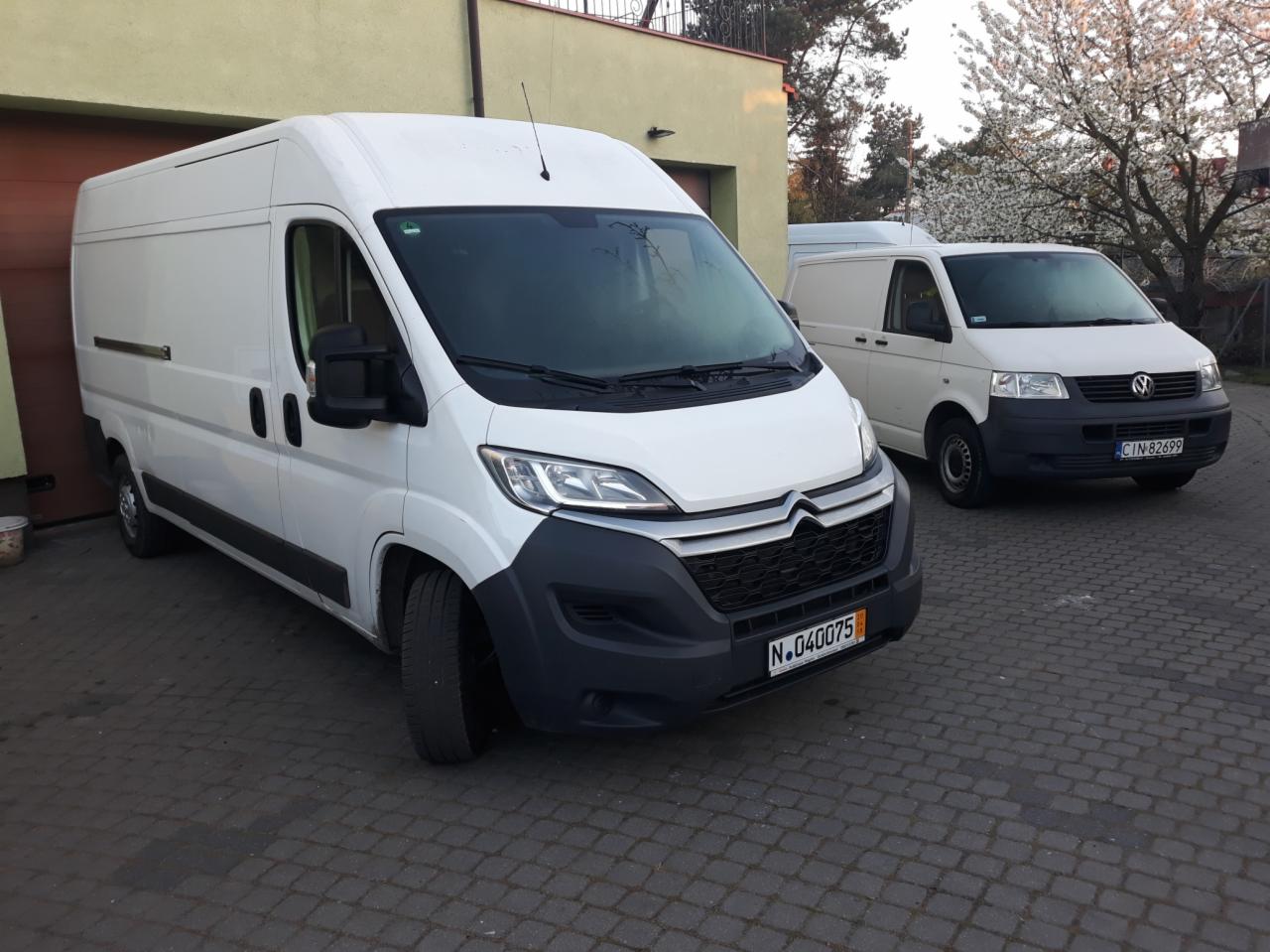 Dwa białe busy zaparkowane na brukowanym podjeździe przed budynkiem z garażem; pierwszy plan pokazuje Citroena Jumper z czarnym zderzakiem i tablicą rejestracyjną N.040075, w tle Volkswagen...
