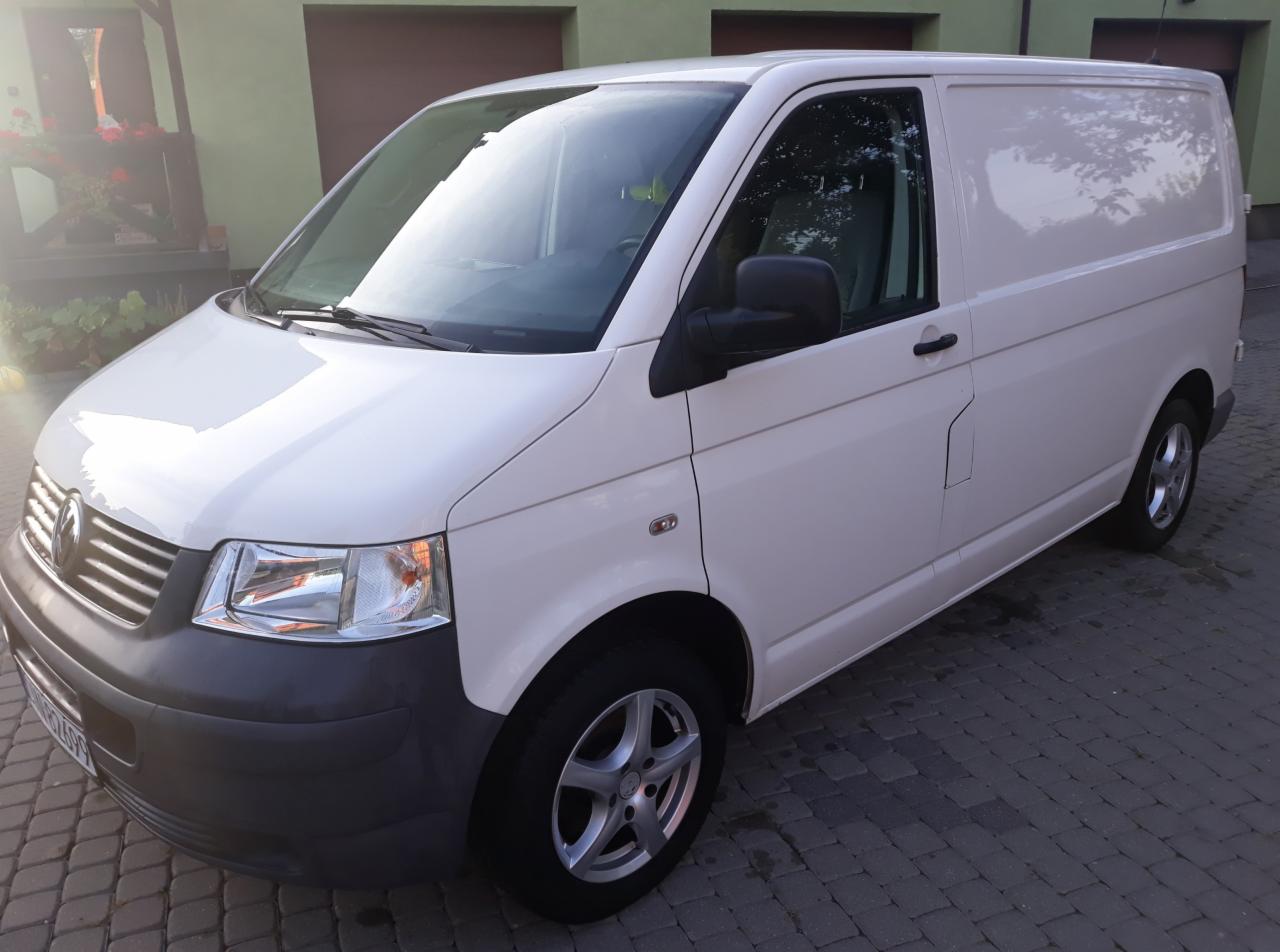 Biały samochód dostawczy Volkswagen Transporter T5 zaparkowany na brukowanym podjeździe przed zielonym budynkiem z brązowymi bramami garażowymi.