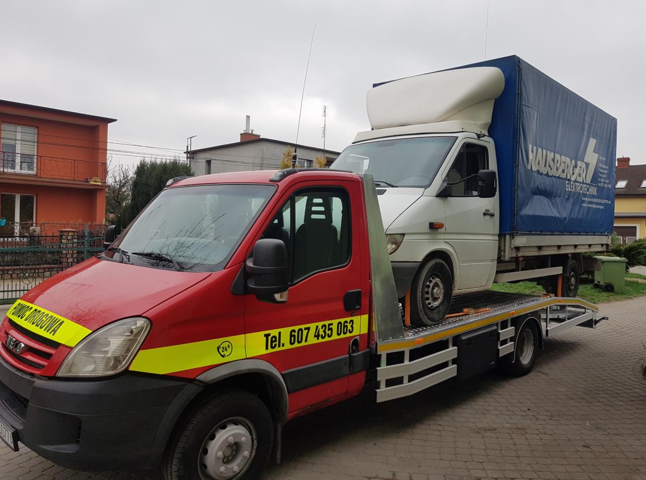 Czerwona laweta z żółtymi pasami i numerem telefonu przewozi białego busa z niebieską plandeką firmy HAUSBERGER ELEKTROTECHNIK.