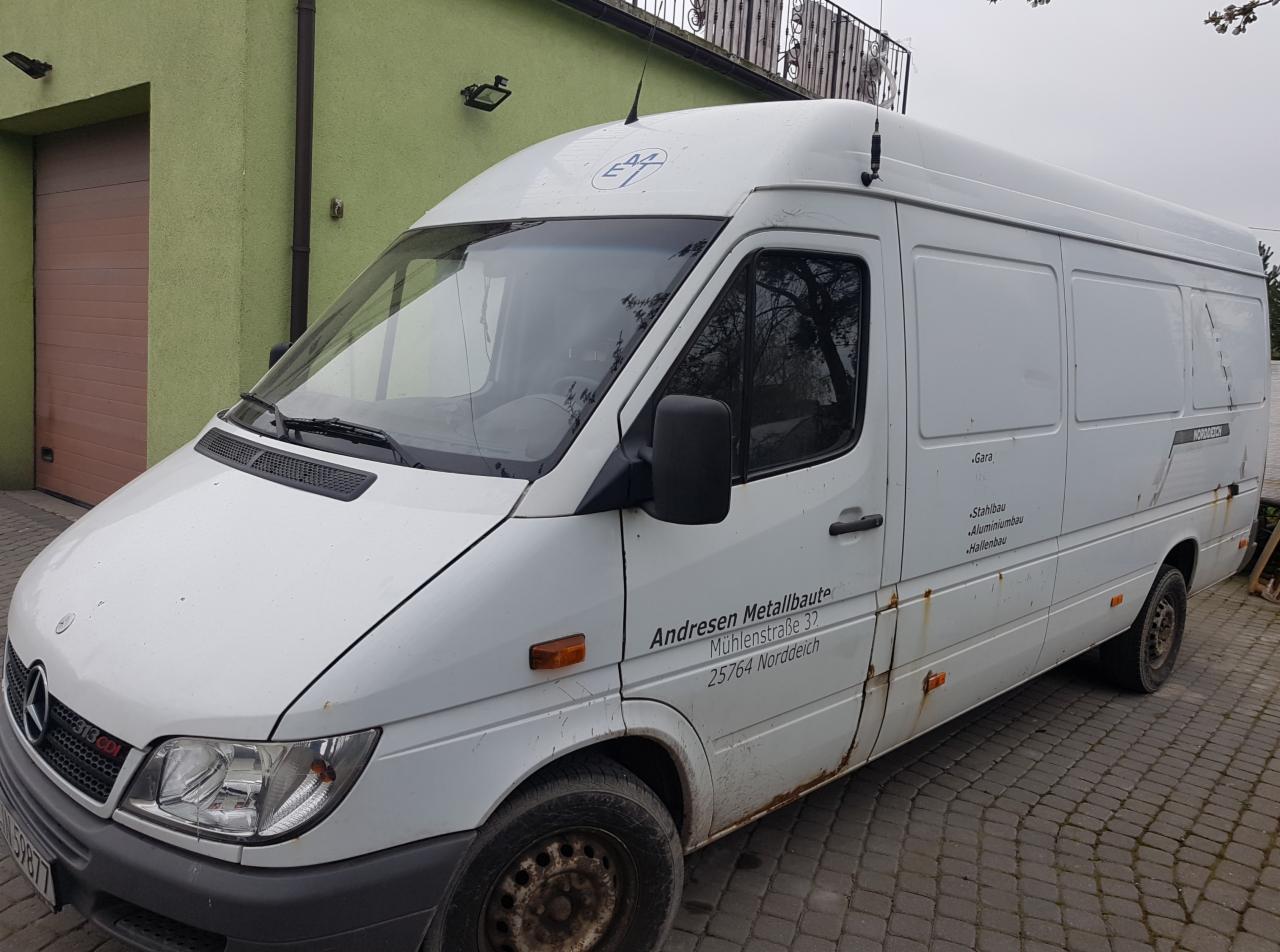 Biały bus Mercedes-Benz Sprinter z widocznymi śladami rdzy na karoserii, zaparkowany na brukowanej nawierzchni przed zielonym budynkiem z brązową bramą garażową. Na boku busa widnieje nazwa firmy...