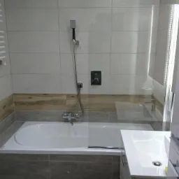 Nowoczesna łazienka z wanną obudowaną szarymi płytkami, szklanym panelem prysznicowym i białym grzejnikiem drabinkowym na ścianie wyłożonej jasnymi płytkami z drewnopodobnym pasem.