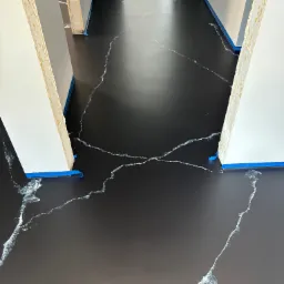 Czarna podłoga z białymi żyłkami imitującymi marmur, zabezpieczona niebieską taśmą malarską przy ścianach i futrynach, widok z góry na korytarz w trakcie wykańczania.