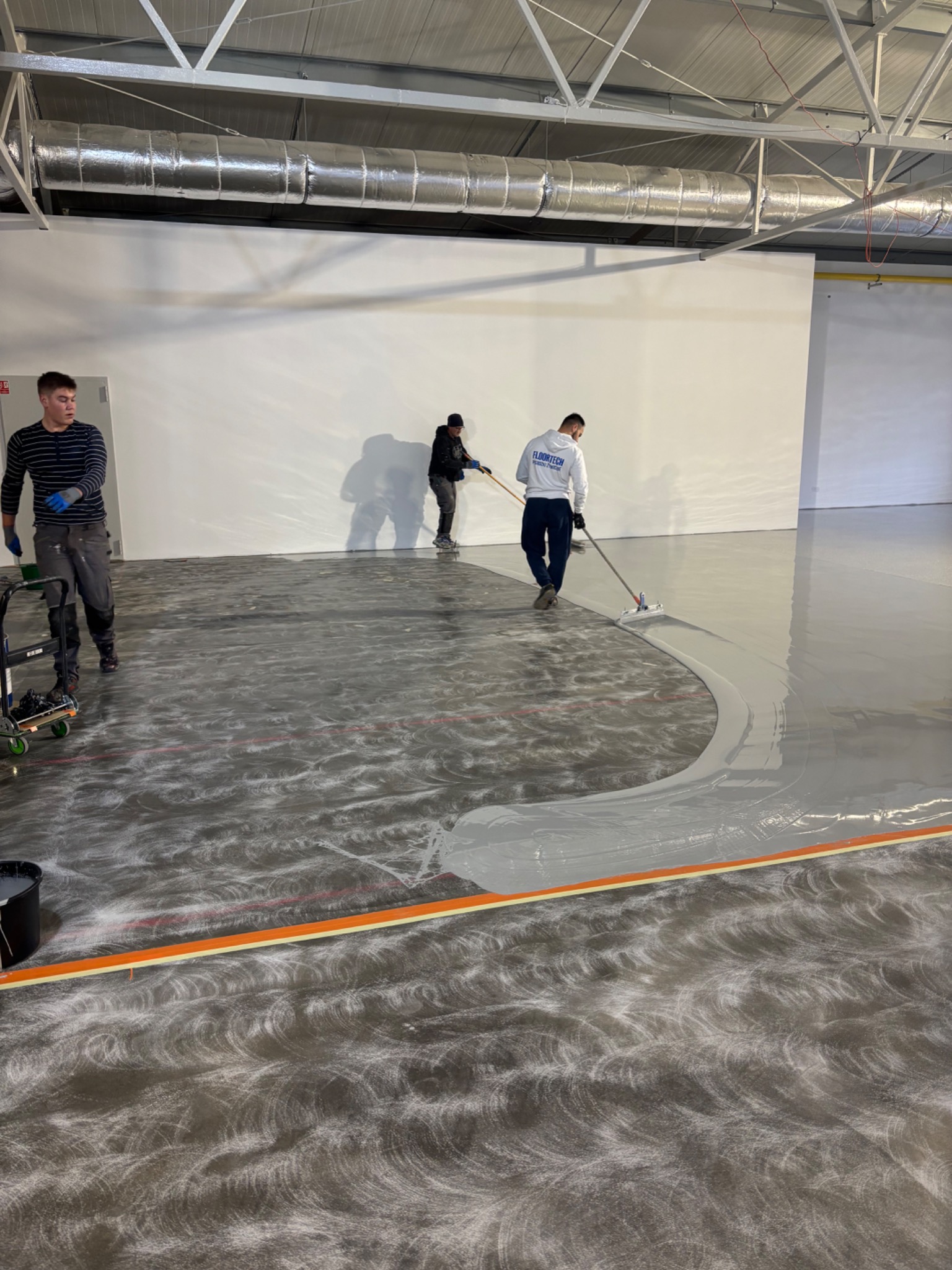 Aplikacja szarej wylewki na posadzce w hali przemysłowej; widoczni trzej pracownicy i narzędzia do rozprowadzania materiału.