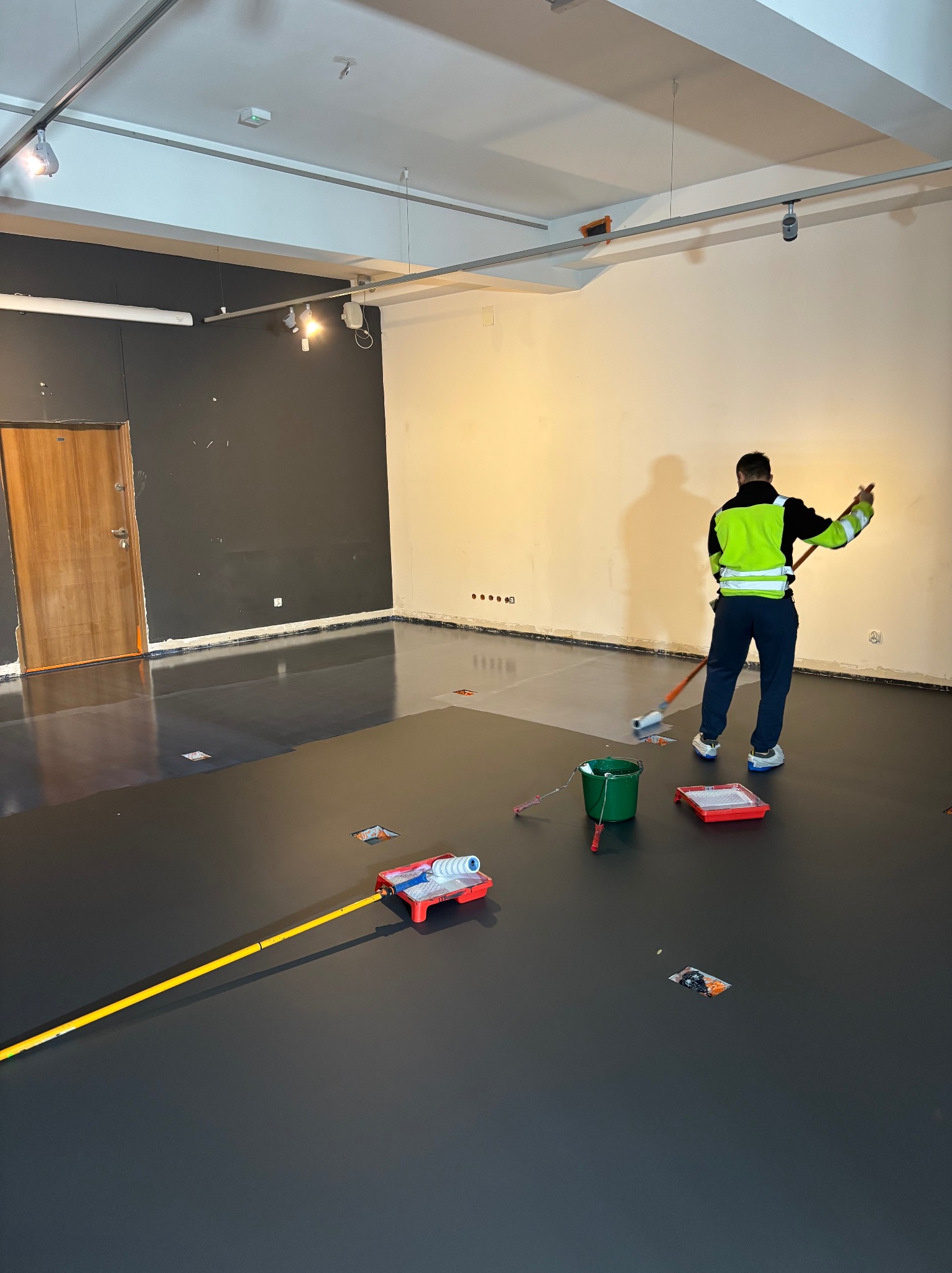 Pracownik w odblaskowej kamizelce aplikuje wałkiem szarą powłokę na posadzce w dużym pomieszczeniu, widoczne narzędzia i akcesoria malarskie, ściany w kolorach szarym i kremowym, oświetlenie sufitowe.