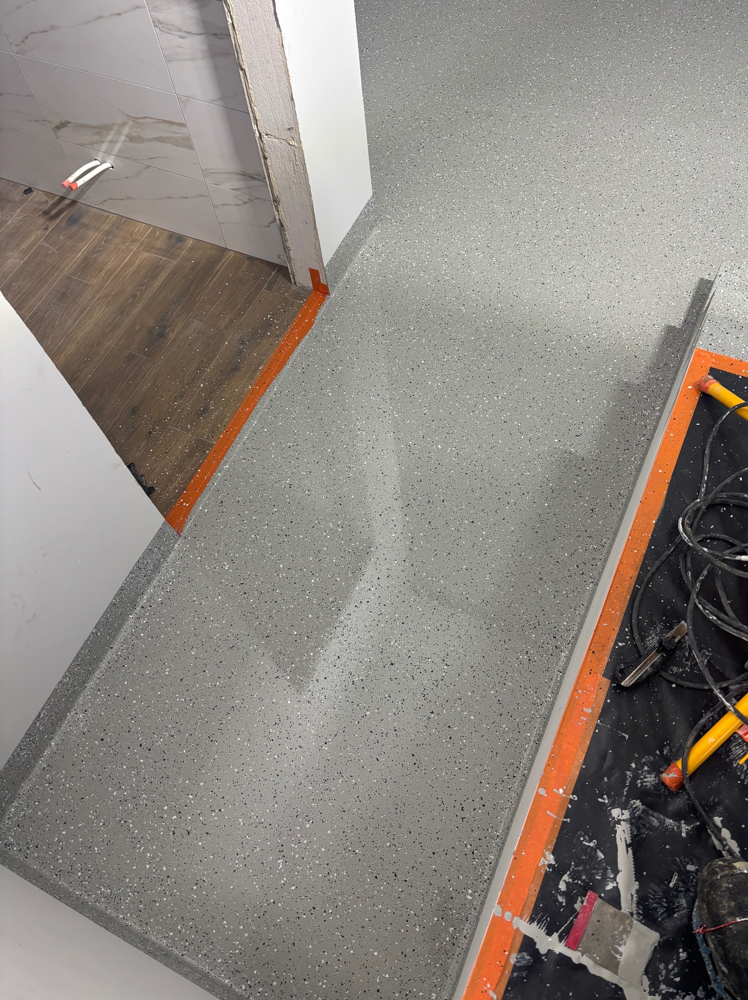 Szara, drobnoziarnista posadzka w jasnym pomieszczeniu, z widocznymi śladami prac wykończeniowych przy ścianie i narzędziami na folii ochronnej.