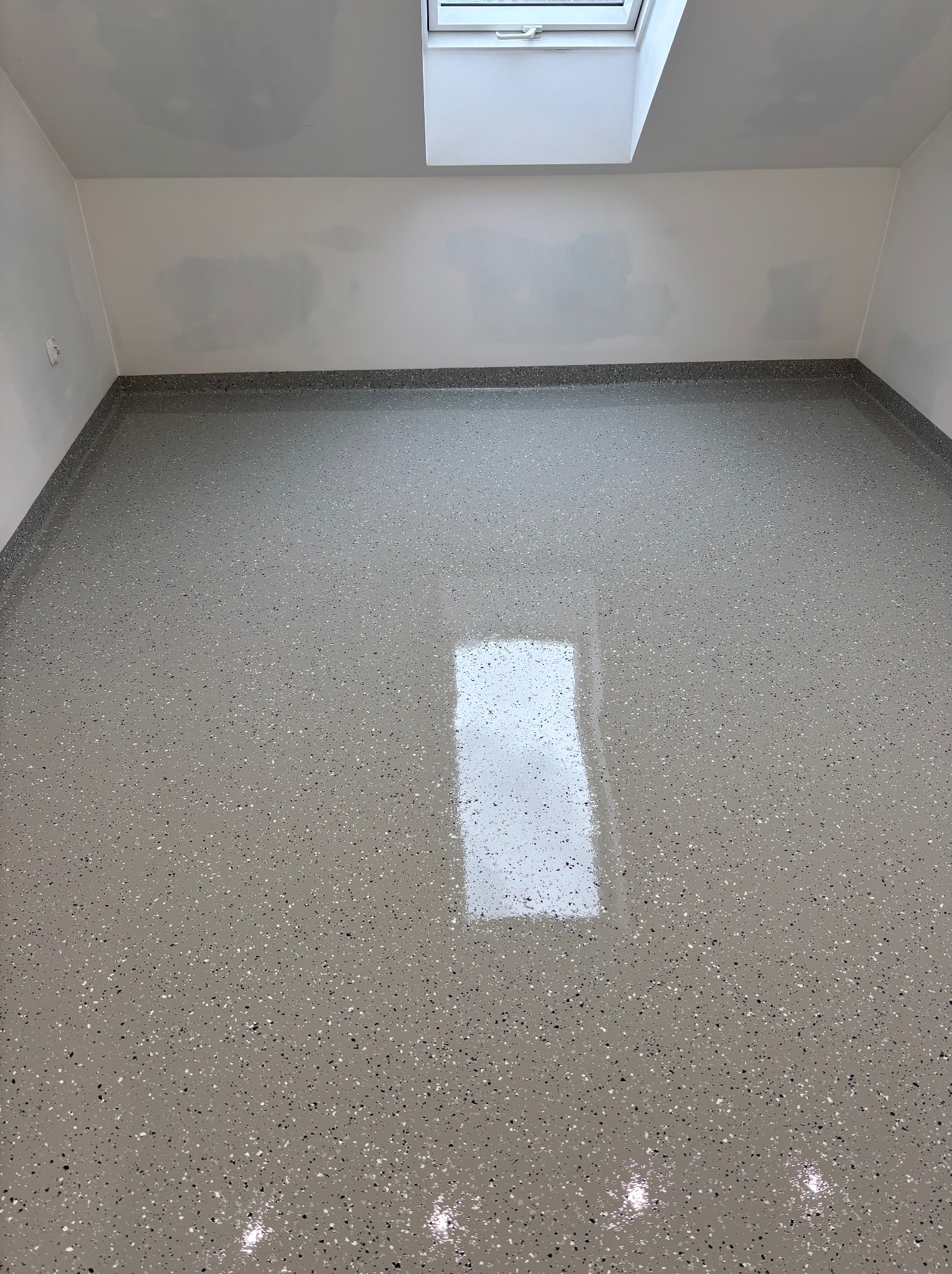 Jasnoszara, błyszcząca podłoga z drobnymi, ciemnymi i jasnymi inkluzjami, odbijająca światło z okna dachowego. Ściany białe, częściowo zagruntowane, z widocznymi śladami po malowaniu.