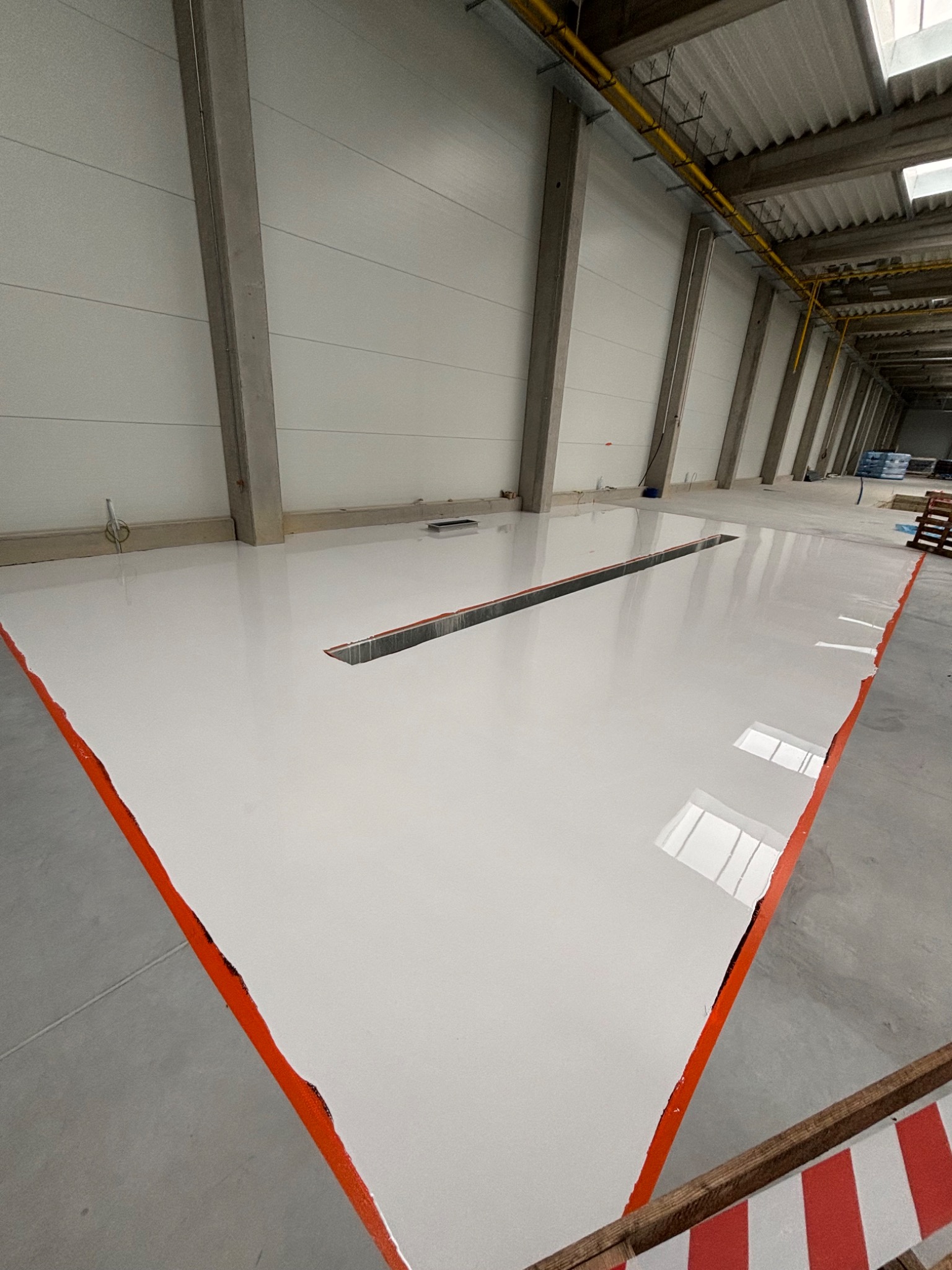 Biała, gładka posadzka w hali przemysłowej z widocznym odpływem liniowym i pomarańczową taśmą wyznaczającą granice powierzchni. W tle widoczne słupy konstrukcyjne i instalacje pod sufitem.