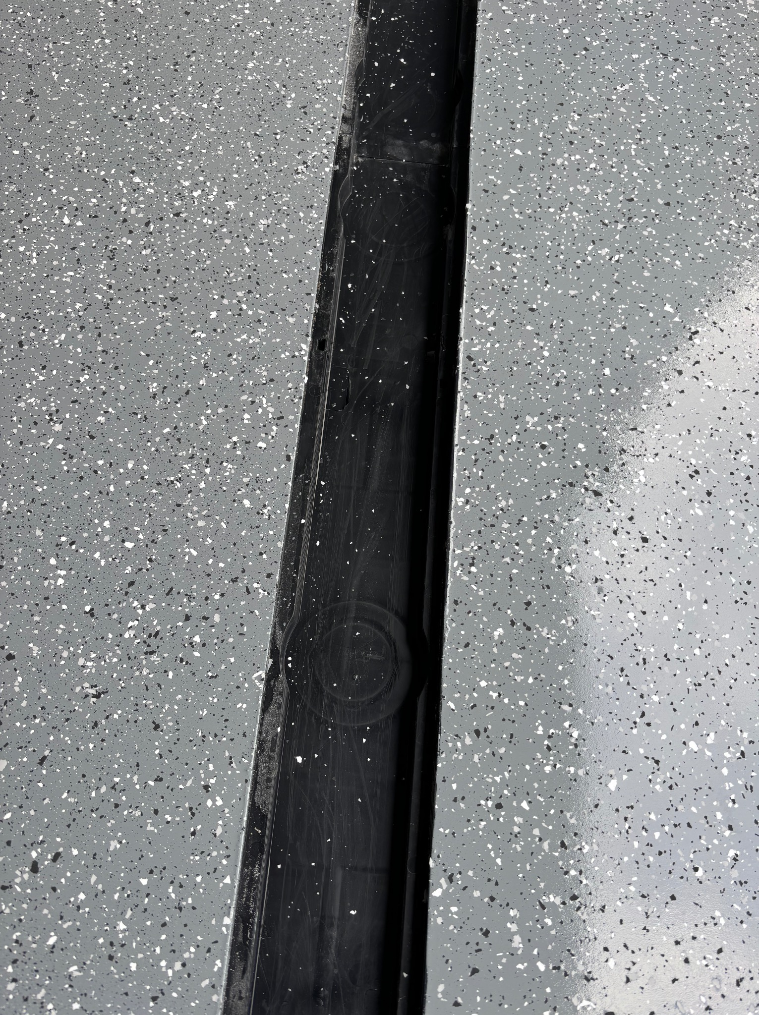 Szara powierzchnia podłogi z czarnymi i białymi plamkami, oddzielona czarnym, podłużnym odpływem liniowym.