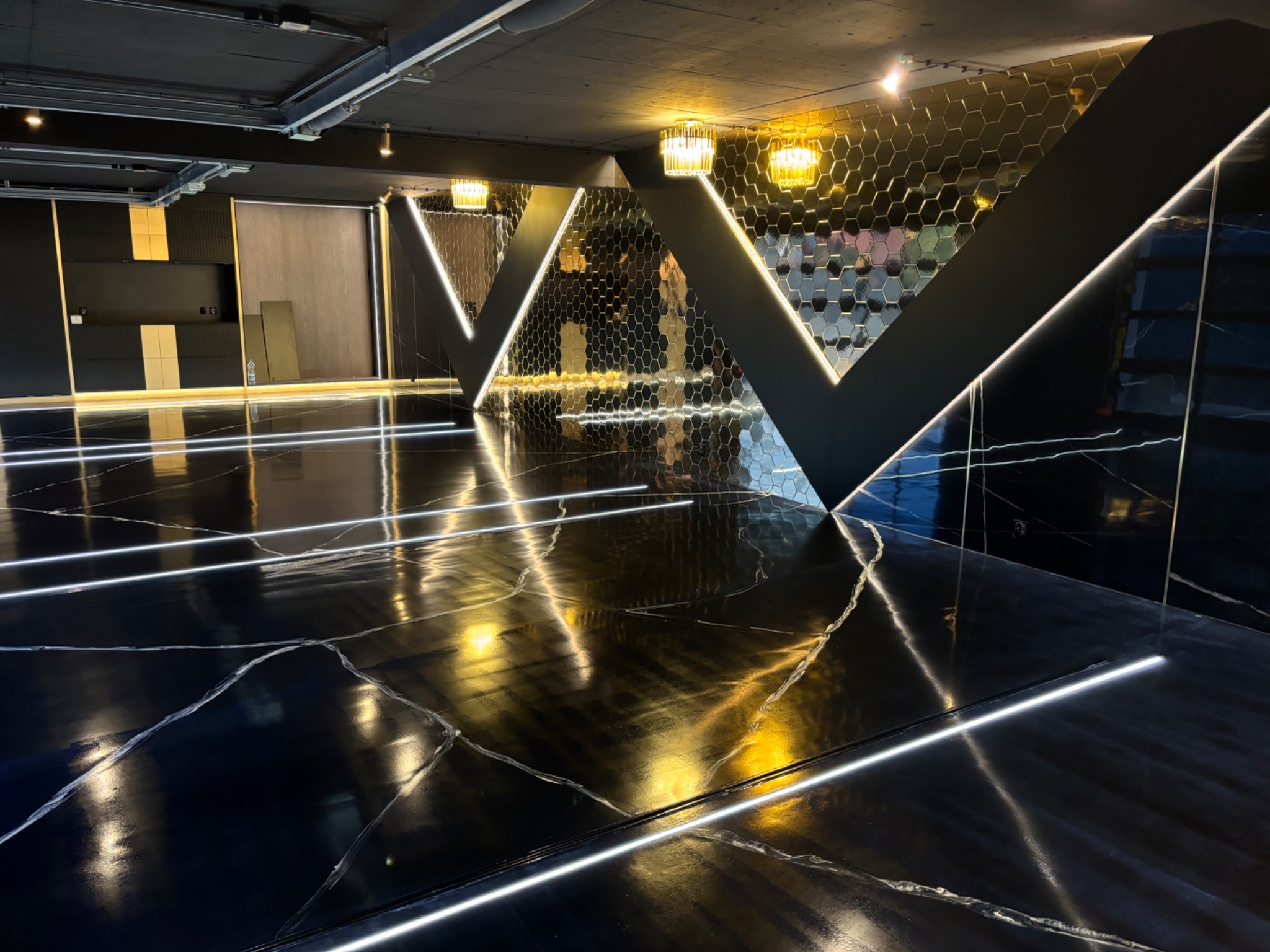 Błyszcząca, czarna podłoga z białymi żyłkami imitującymi marmur, oświetlona liniowymi lampami LED, z dekoracyjnymi ścianami z heksagonalnych płytek i geometrycznymi elementami.
