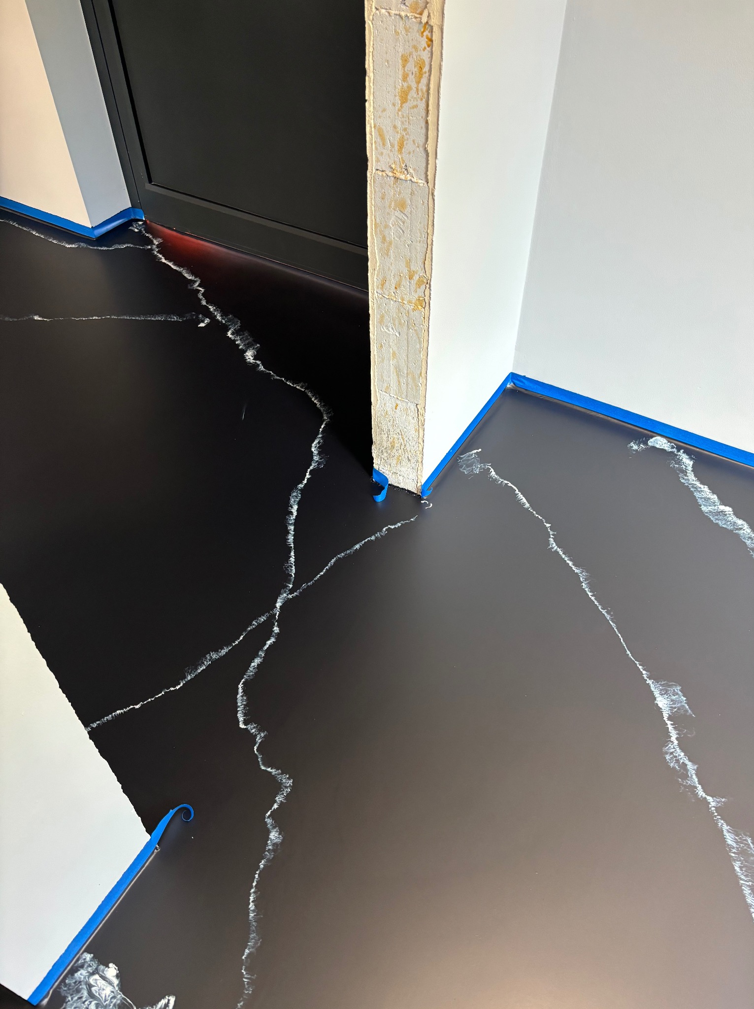 Czarna podłoga z białymi żyłkami imitującymi marmur, zabezpieczona niebieską taśmą malarską przy ścianach i ościeżnicy przed aplikacją wykończenia.