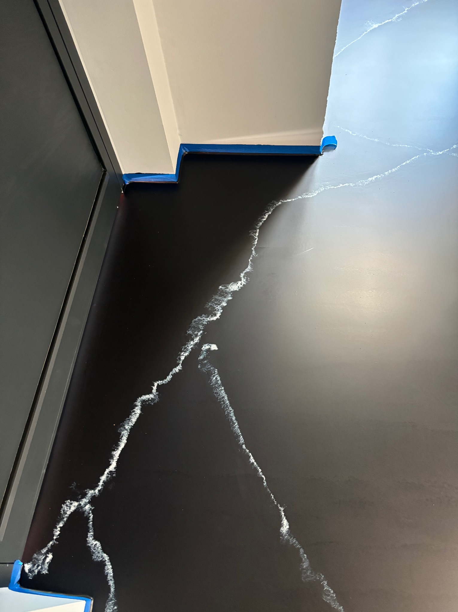 Czarna podłoga z efektem żyłkowania, zabezpieczona niebieską taśmą malarską przy ścianach i framugach drzwi, w trakcie prac wykończeniowych.