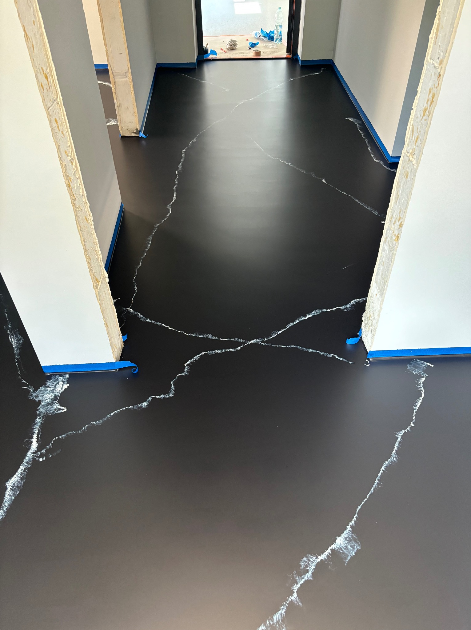 Czarna podłoga z białymi żyłkami imitującymi marmur, zabezpieczona niebieską taśmą malarską przy ścianach i futrynach, widok z góry na korytarz w trakcie wykańczania.