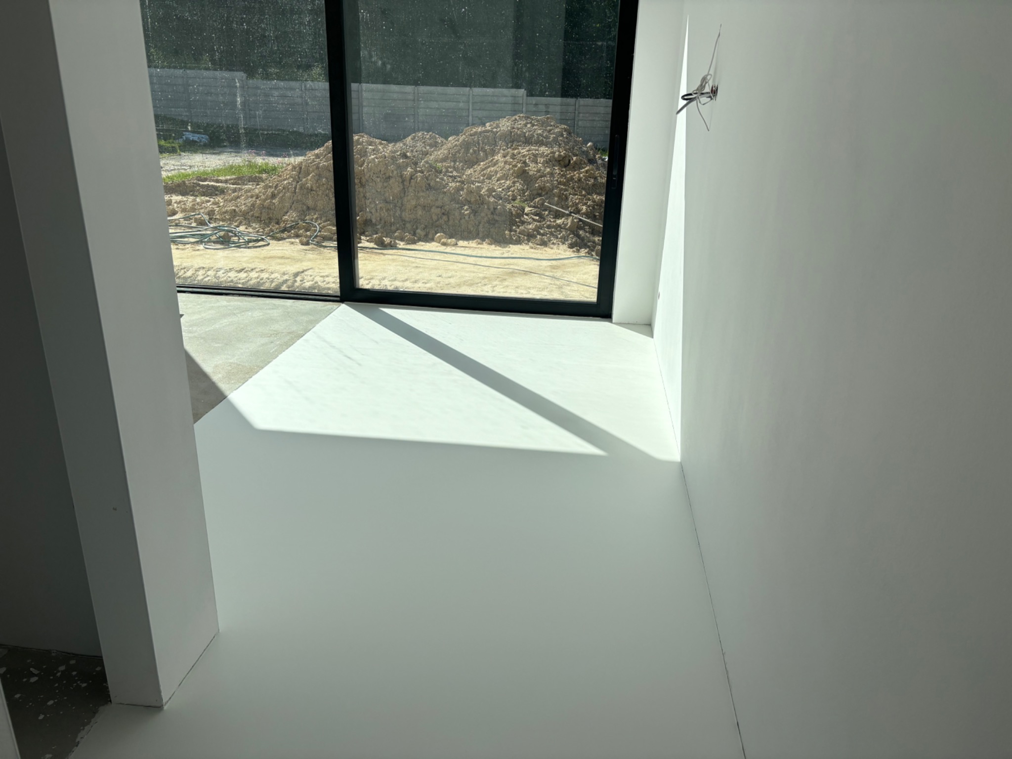 Świeżo wylana, jasnoszara posadzka żywiczna w surowym wnętrzu z widokiem na stertę ziemi przez duże okno.