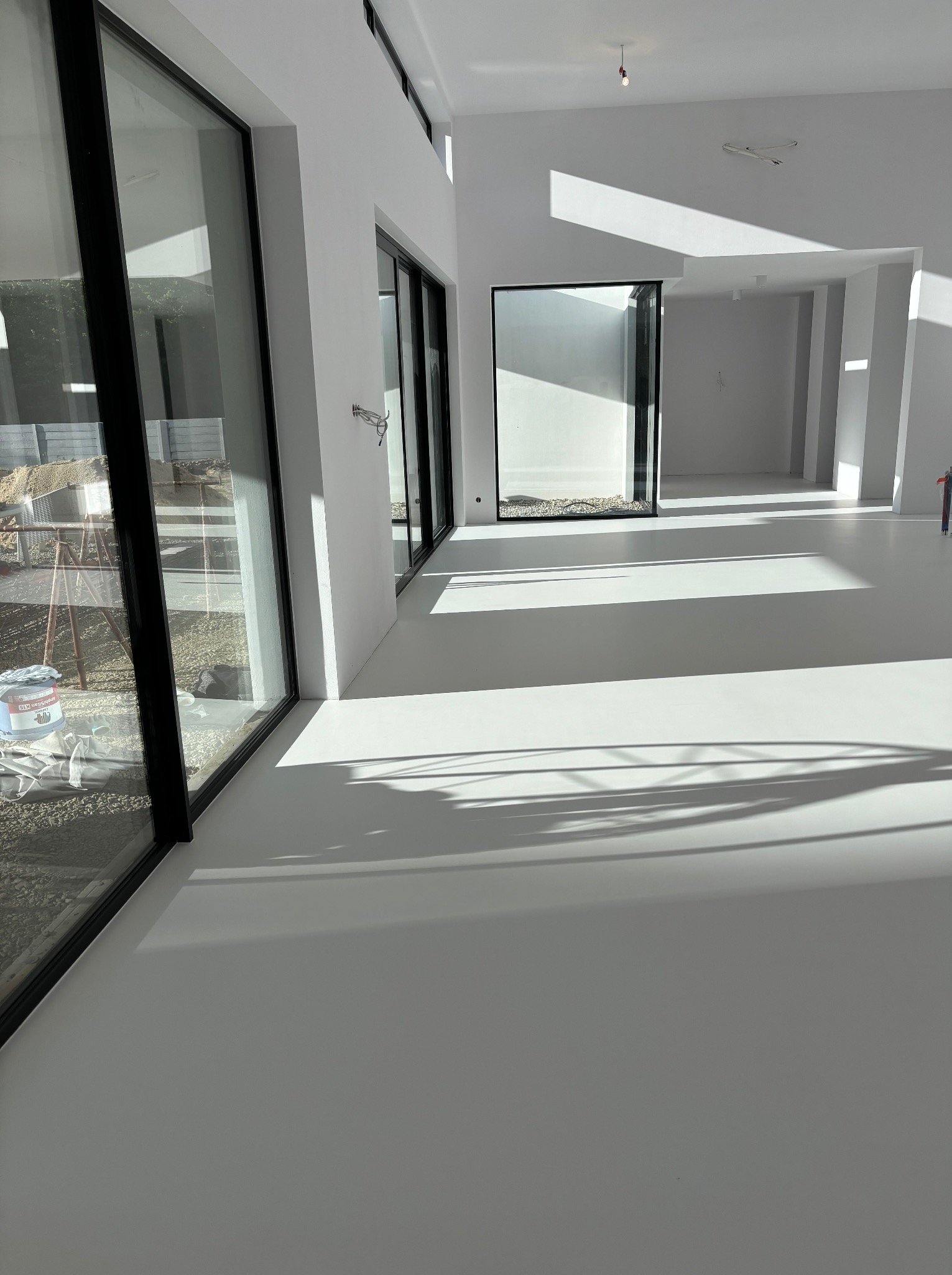Jasne, przestronne wnętrze z białą, jednolitą posadzką i dużymi oknami. Na zewnątrz widoczne materiały budowlane i wiadro z farbą. Cienie padające przez okna tworzą geometryczne wzory na podłodze.