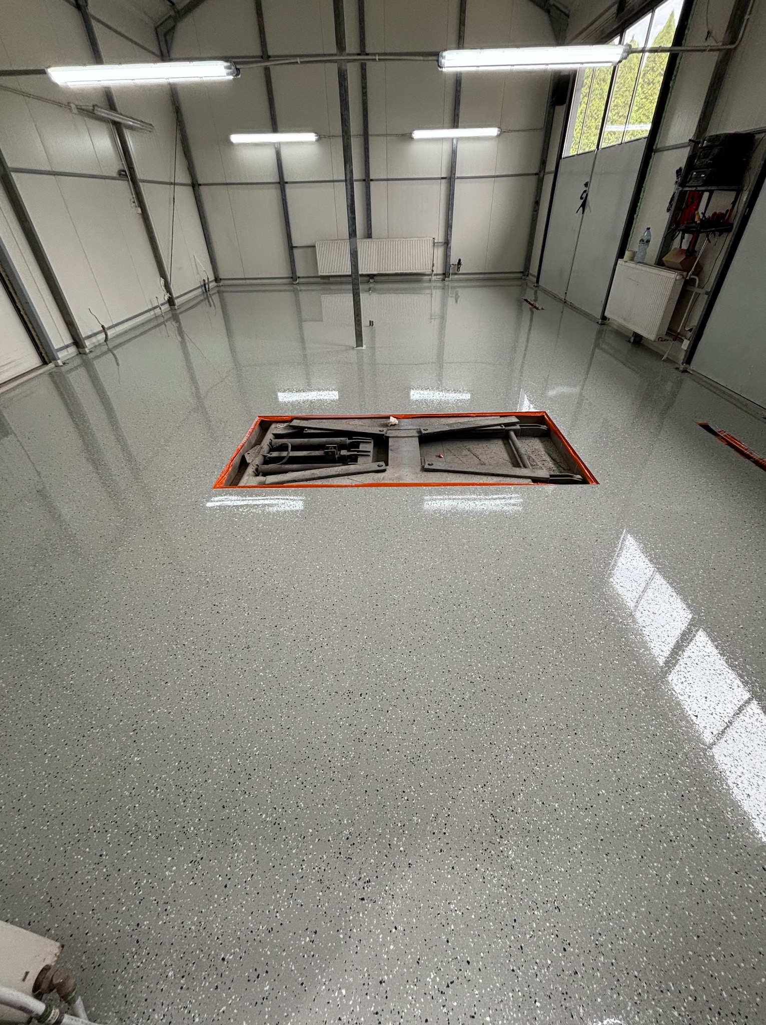 Jasnoszara, błyszcząca posadzka z drobnymi plamkami w hali przemysłowej, z widocznym kanałem rewizyjnym z pomarańczową ramą i podnośnikiem wewnątrz.