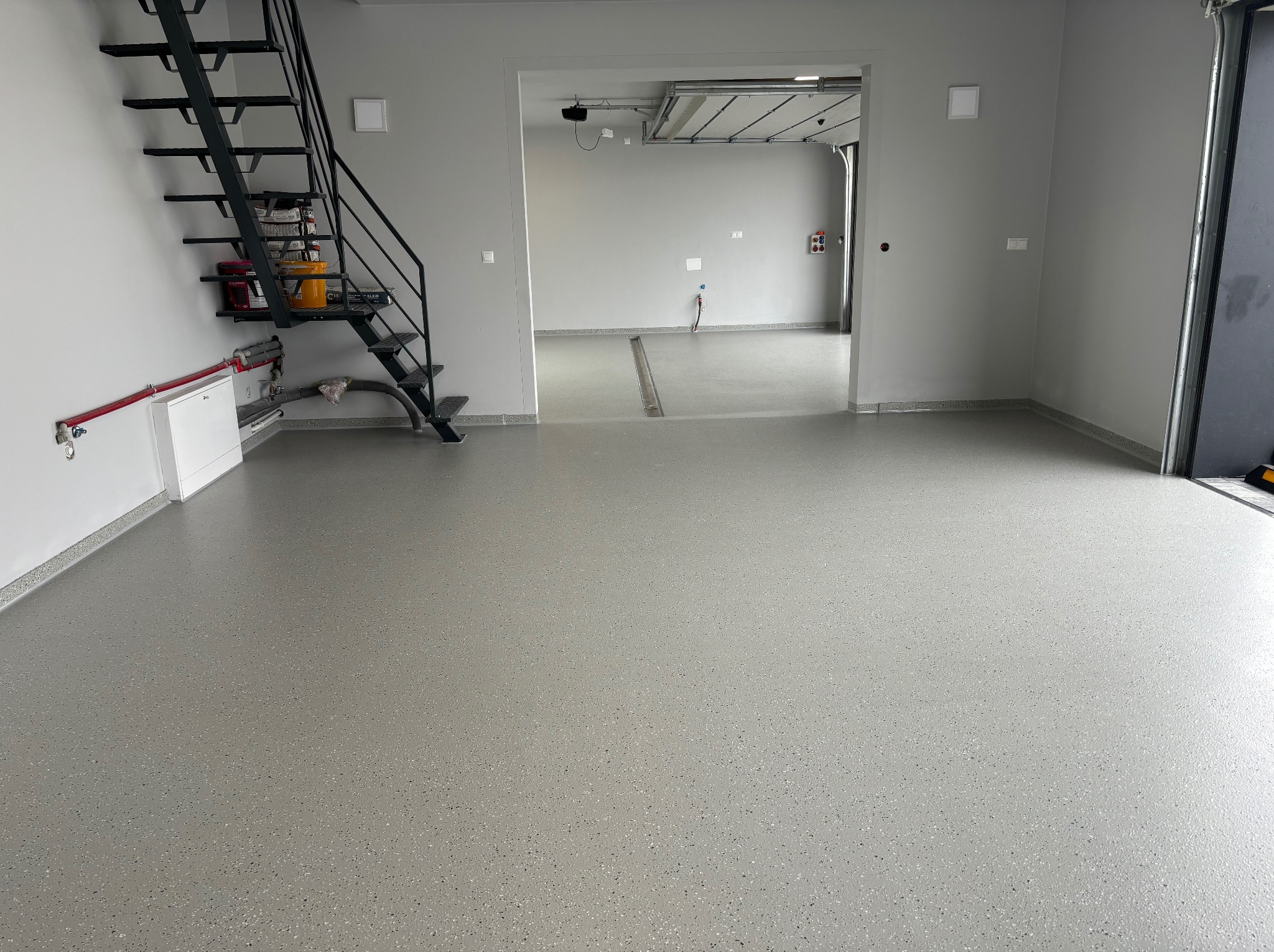 Jasnoszara, gładka posadzka z delikatnym wzorem w garażu lub hali, widoczne schody metalowe i otwarty wjazd do drugiego pomieszczenia.