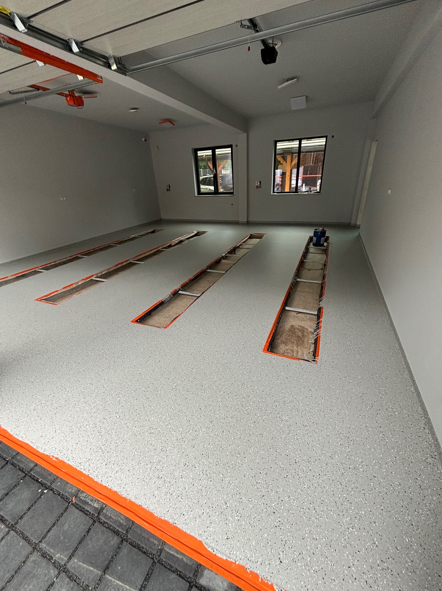 Jasnoszara posadzka w garażu z widocznymi otworami kanałów naprawczych, obramowanymi pomarańczową farbą, z dodatkiem kolorowych drobinek w strukturze, oświetlona światłem dziennym wpadającym przez...