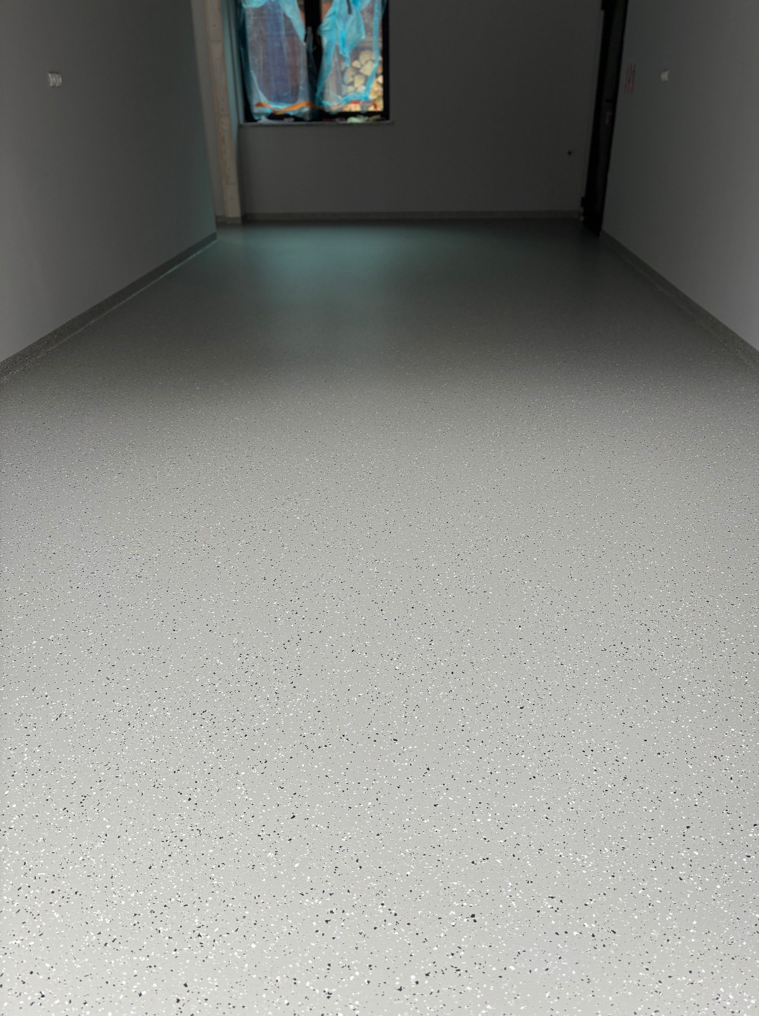 Jasnoszara posadzka z drobnymi czarnymi i białymi inkluzjami, świeżo wylana w pomieszczeniu z szarymi ścianami i oknem zabezpieczonym folią.