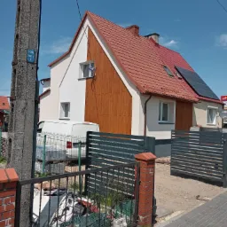 Nowoczesny dom jednorodzinny z częściowo odnowioną elewacją, gdzie jedna strona jest biała, a druga wykończona drewnopodobnymi panelami pionowymi; czerwony dach z panelami słonecznymi, widoczny zza...