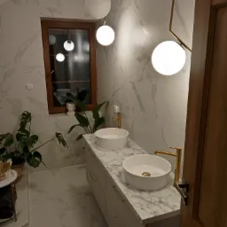 Elegancka łazienka z dwoma białymi umywalkami nablatowymi na blacie z imitacją marmuru, złotymi bateriami, marmurowymi płytkami na ścianach i podłodze, oświetleniem w postaci kulistych lamp...