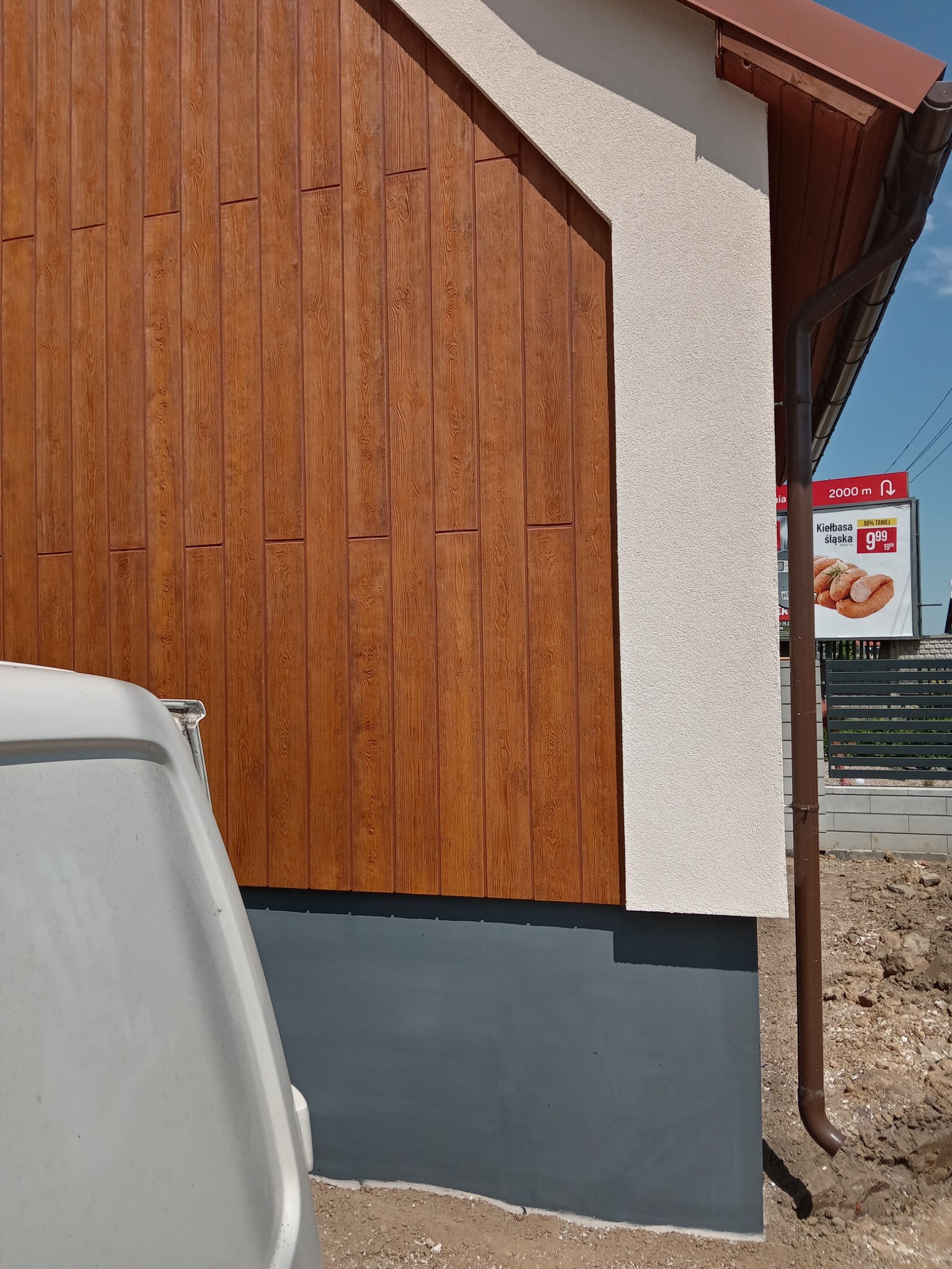 Fragment elewacji budynku z brązową okładziną imitującą drewno, połączonej z jasnym tynkiem strukturalnym i szarą podmurówką. Widoczny fragment białego samochodu dostawczego oraz rynna. W tle...