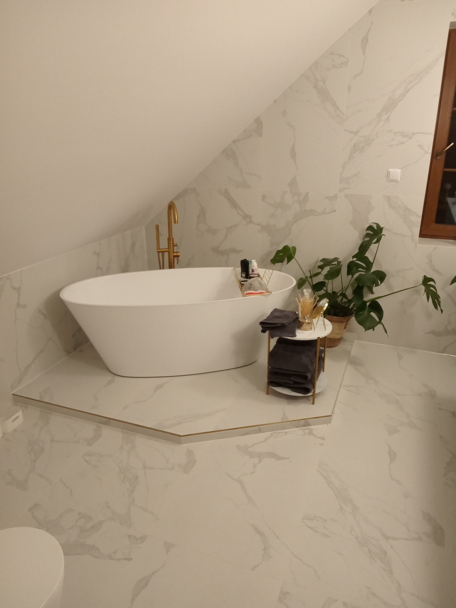 Nowoczesna łazienka na poddaszu z białą wanną wolnostojącą, marmurowymi płytkami na ścianach i podłodze, złotą armaturą, rośliną doniczkową Monstera i stolikiem z ręcznikami.