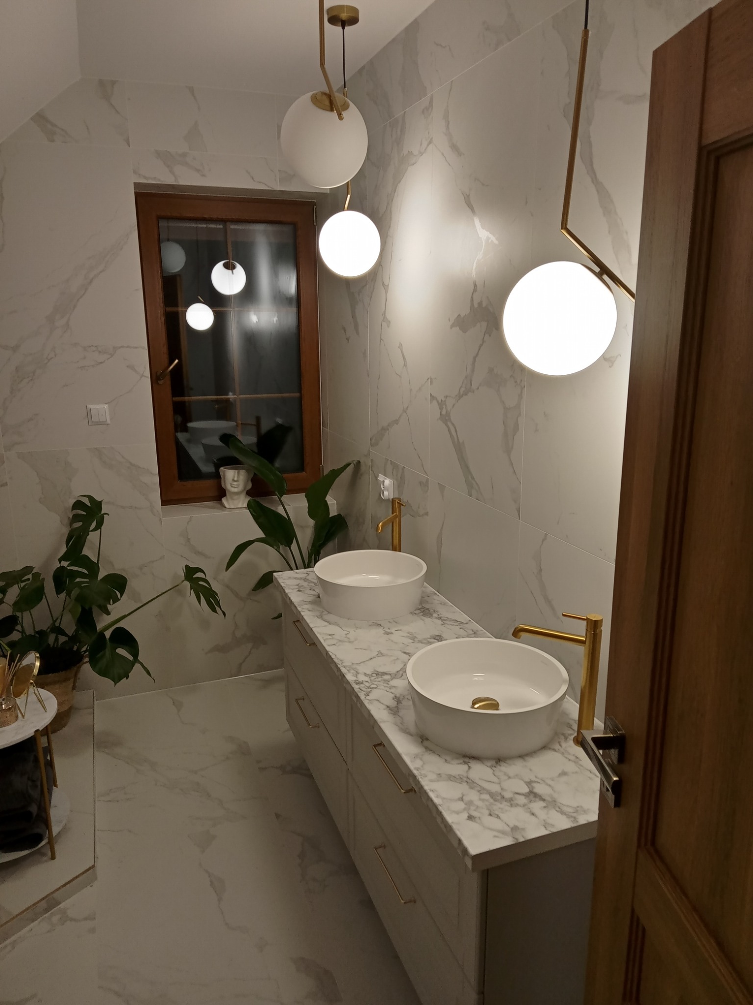 Elegancka łazienka z dwoma białymi umywalkami nablatowymi na blacie z imitacją marmuru, złotymi bateriami, marmurowymi płytkami na ścianach i podłodze, oświetleniem w postaci kulistych lamp...