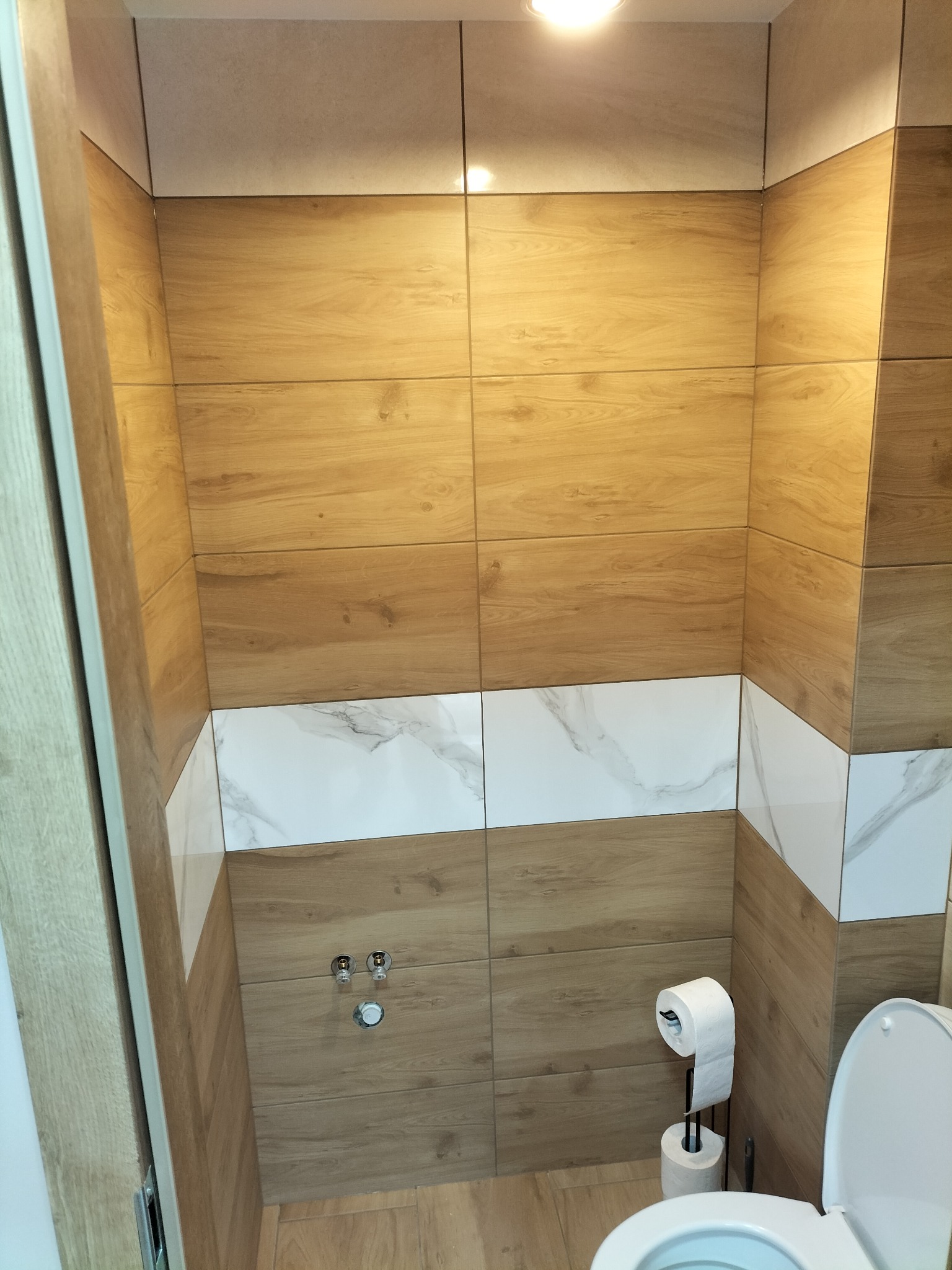 Wykończona łazienka z drewnopodobnymi i marmurowymi płytkami na ścianach, widoczna toaleta z otwartą klapą i uchwyt na papier toaletowy z dwoma rolkami, armatura na ścianie.