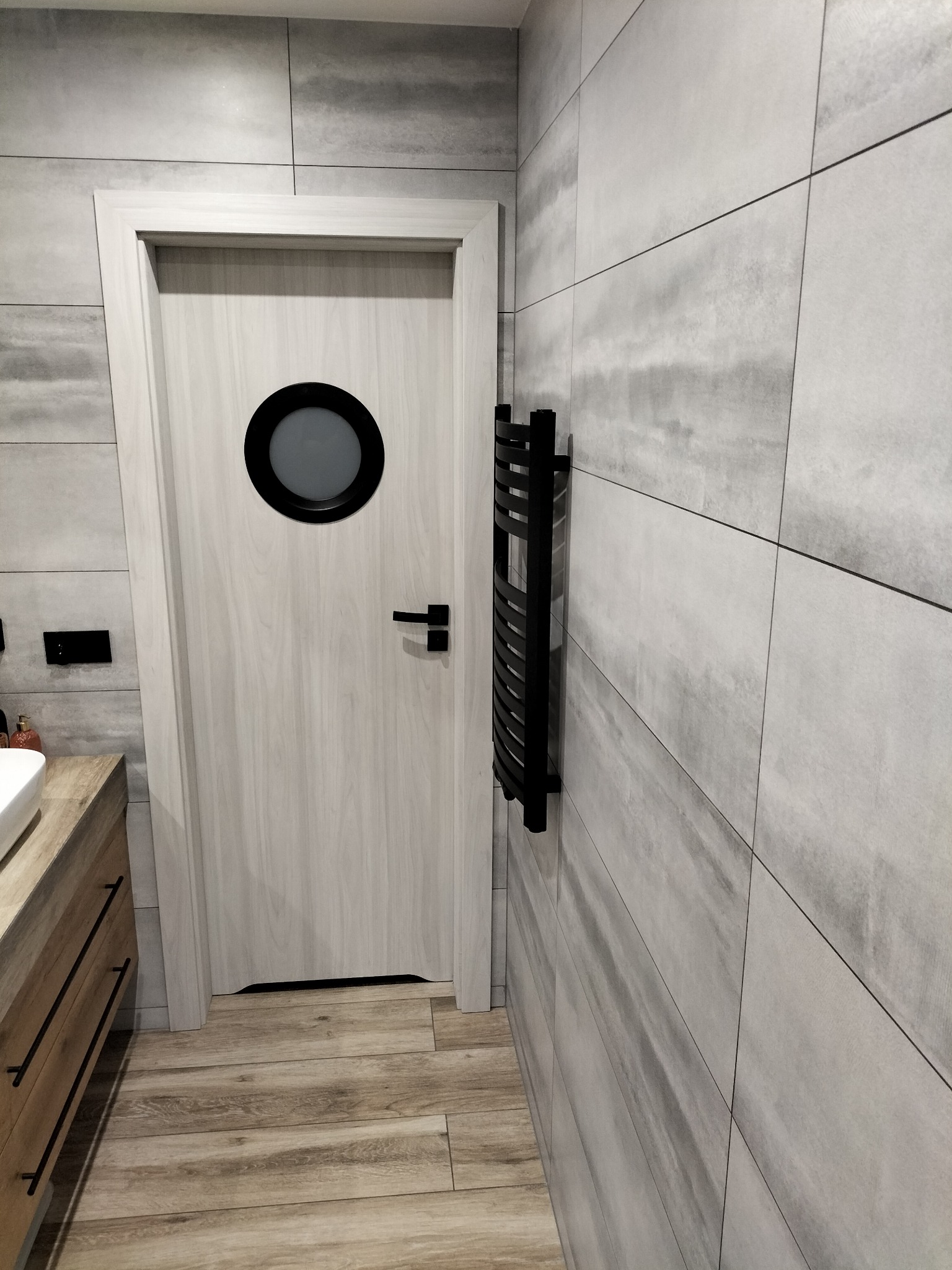 Nowoczesna łazienka z szarymi płytkami imitującymi beton, drewnopodobną podłogą, białą umywalką z szafką i drzwiami w stylu marynistycznym z okrągłym oknem oraz czarnym grzejnikiem drabinkowym.