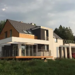 Nowoczesny dom jednorodzinny z elementami drewnianymi i grafitowymi, z dużym tarasem i zadaszonym miejscem parkingowym, otoczony zielenią.