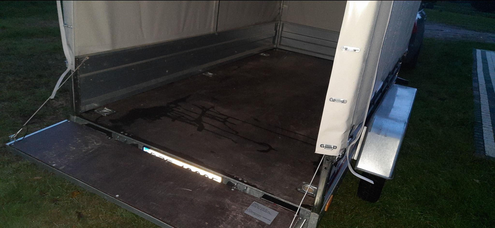 Otwarta przyczepa z plandeką, widoczna pusta przestrzeń ładunkowa z drewnianą podłogą i metalowymi ścianami, oświetlenie odblaskowe na tylnej klapie.