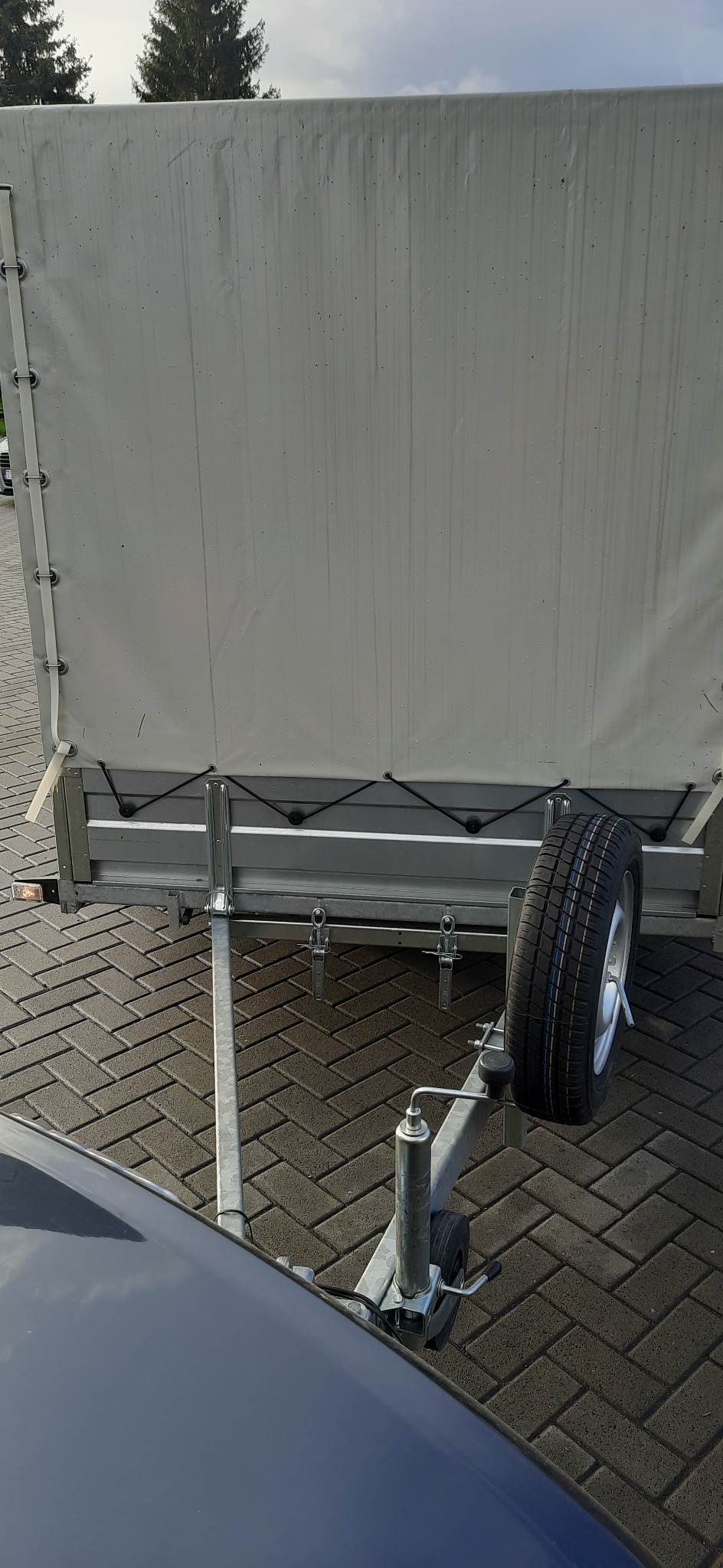 Tylny widok przyczepy z plandeką i kołem zapasowym, zaparkowanej na brukowanym placu, z widocznym fragmentem samochodu.