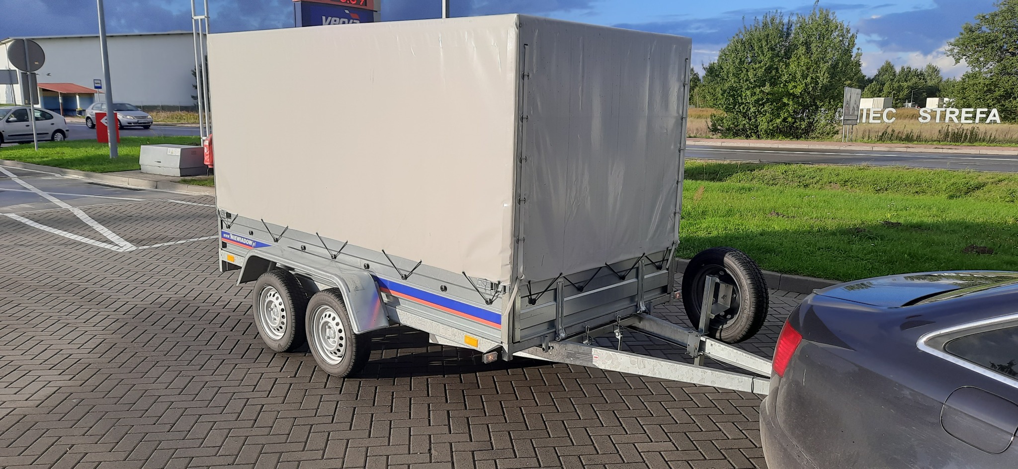 Przyczepa samochodowa z plandeką na parkingu z kostki, widoczny zapasowe koło i fragment samochodu.
