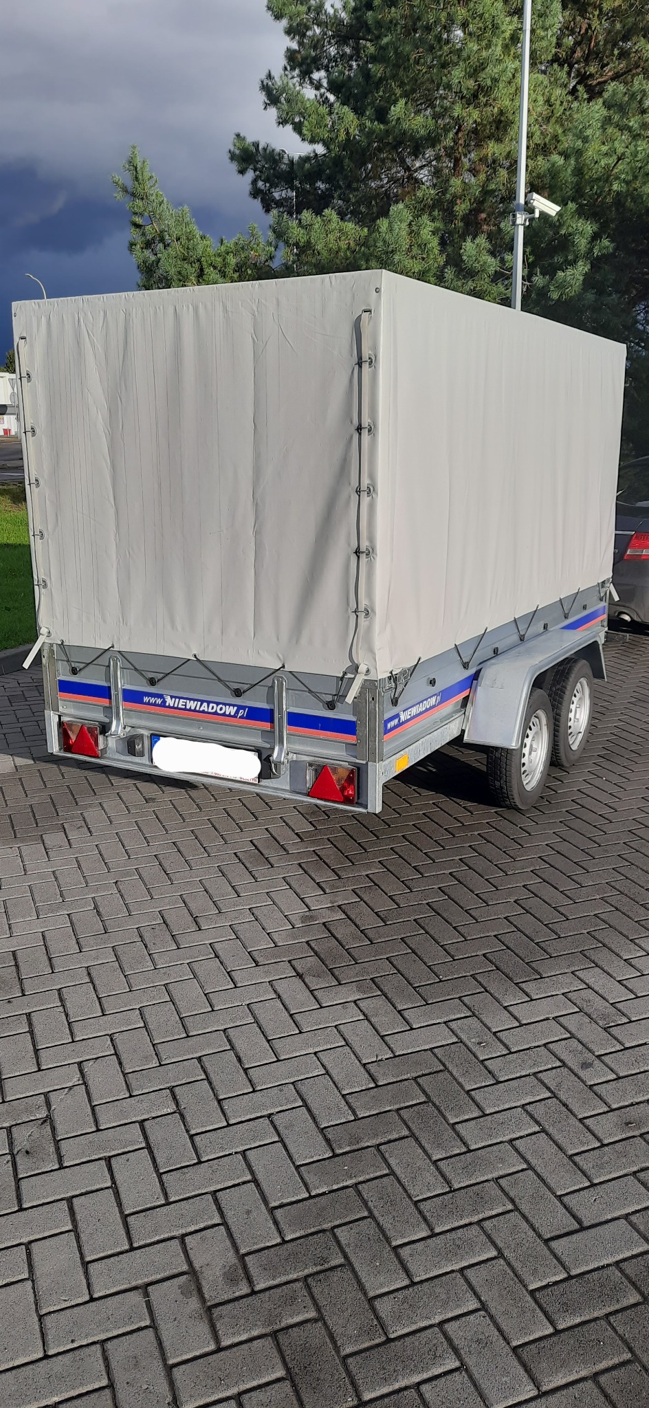 Przyczepa z plandeką marki Niewiadów, widok z tyłu na kostce brukowej, w tle drzewa i pochmurne niebo.