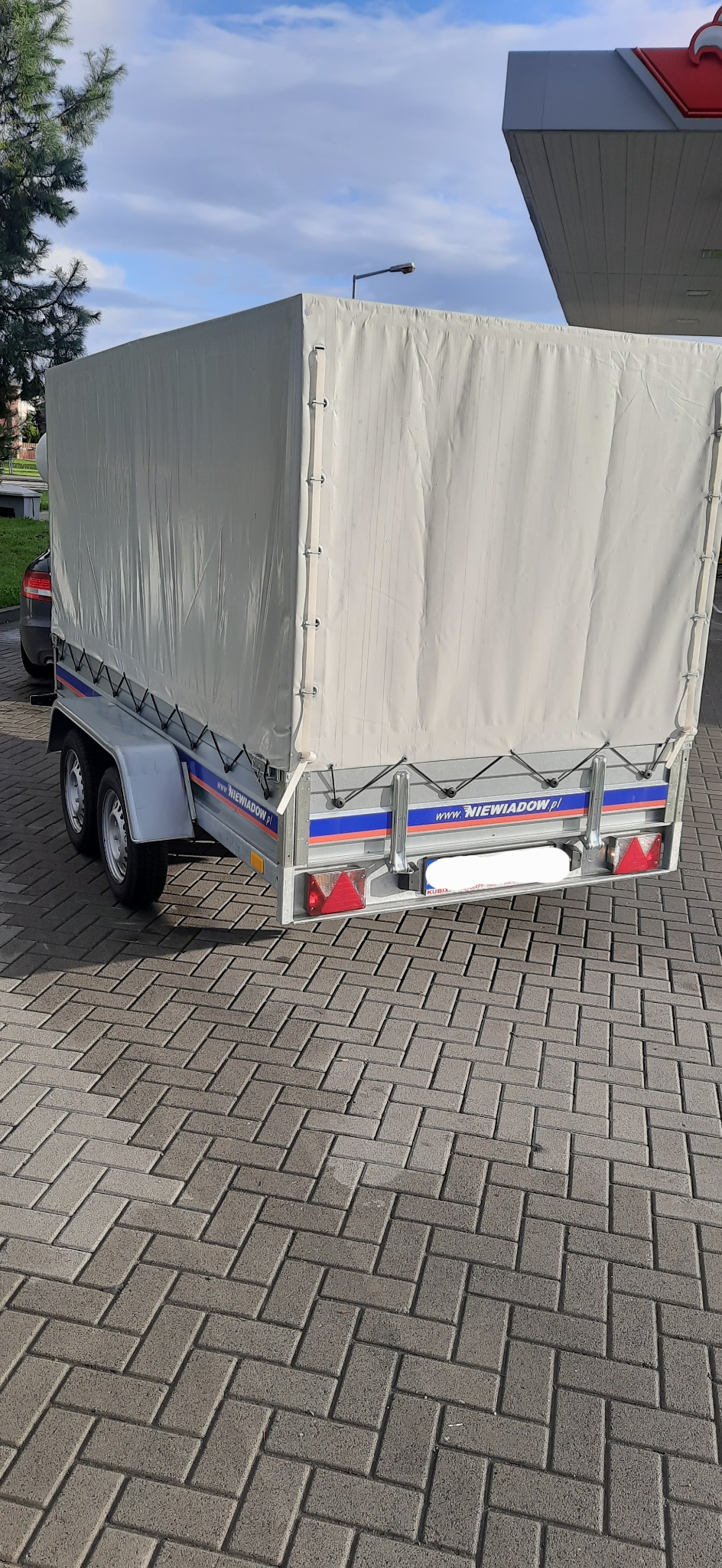 Przyczepa samochodowa z plandeką marki Niewiadów na kostce brukowej, widok z tyłu, z widocznym adresem strony internetowej producenta.