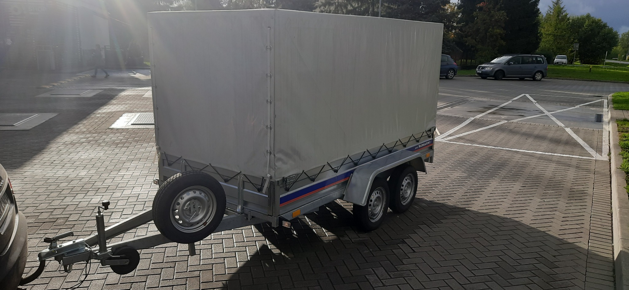 Przyczepa z plandeką, zaparkowana na kostce brukowej, z widocznym kołem zapasowym i hakiem holowniczym.