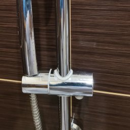 Błysk Firma sprzątająca - Błyszcząca, chromowana bateria prysznicowa z widocznymi kroplami wody na tle ciemnobrązowej ściany z poziomymi paskami. Detal po czyszczeniu.