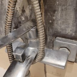 Błysk Firma sprzątająca - Z bliska: zanieczyszczona, chromowana bateria prysznicowa z elastycznym wężem na tle szarej, zabrudzonej ściany. Widoczne osady i zacieki.