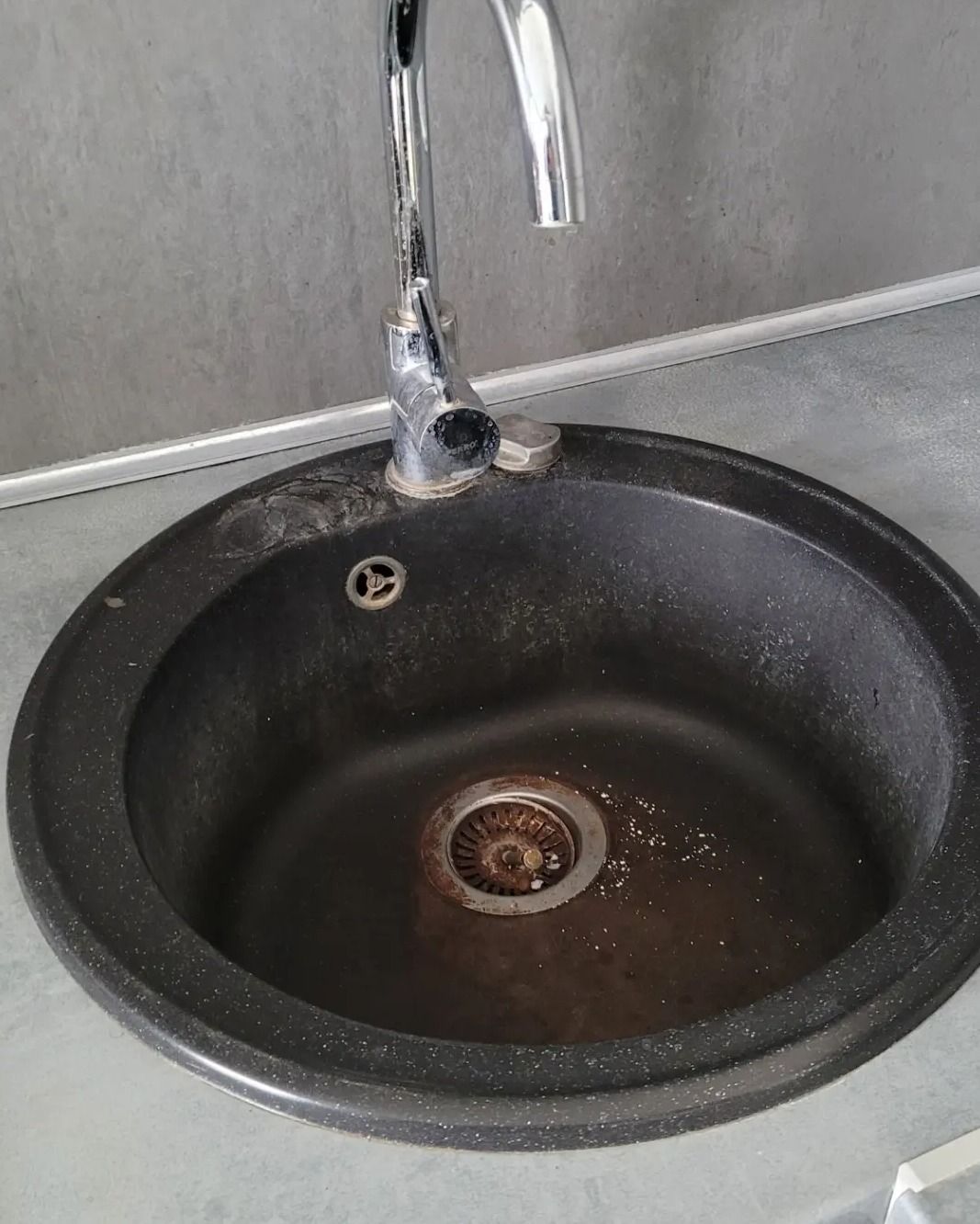 Z bliska: okrągły, czarny zlew z rdzawym odpływem i chromowaną baterią. Widoczne ślady użytkowania i osad na powierzchni zlewu.