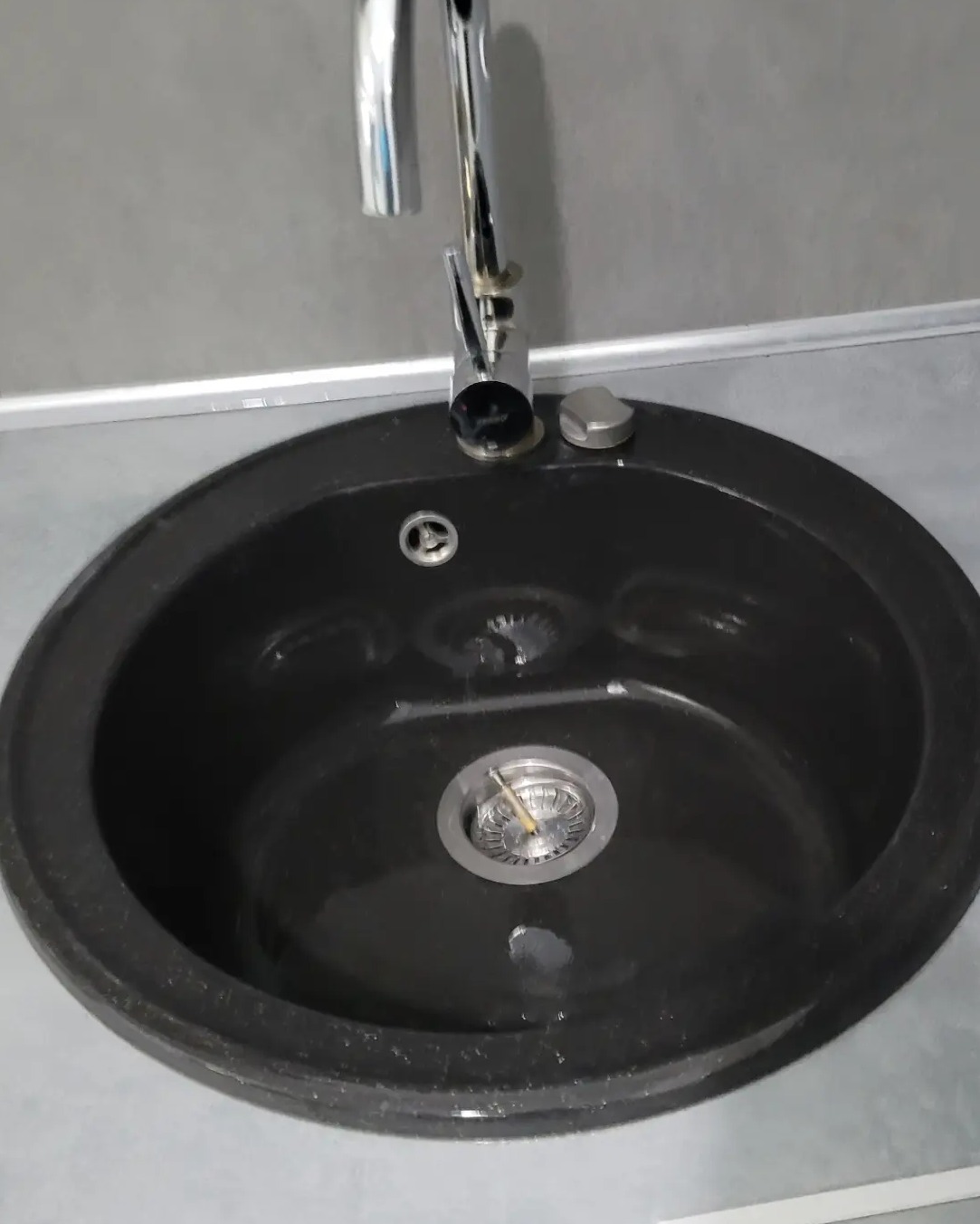 Okrągła, czarna umywalka granitowa z chromowaną baterią, widok z góry. Minimalistyczne wnętrze, szare tło. Czysta powierzchnia zlewu.