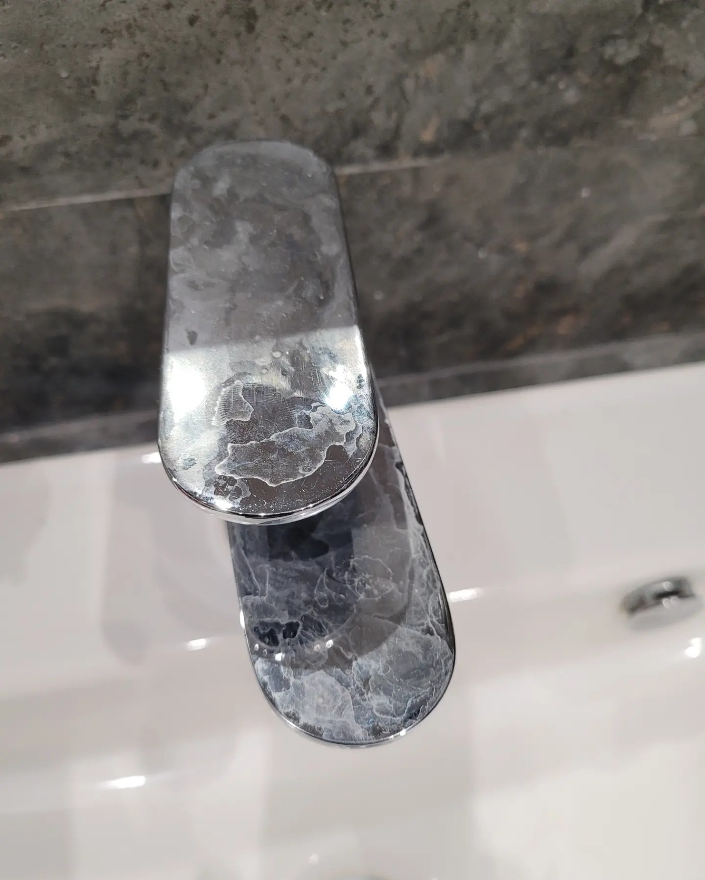 Z bliska: zacieki z kamienia na chromowanej baterii umywalkowej, widoczne na tle białej umywalki i szarej ściany. Nierównomierne osady mineralne.