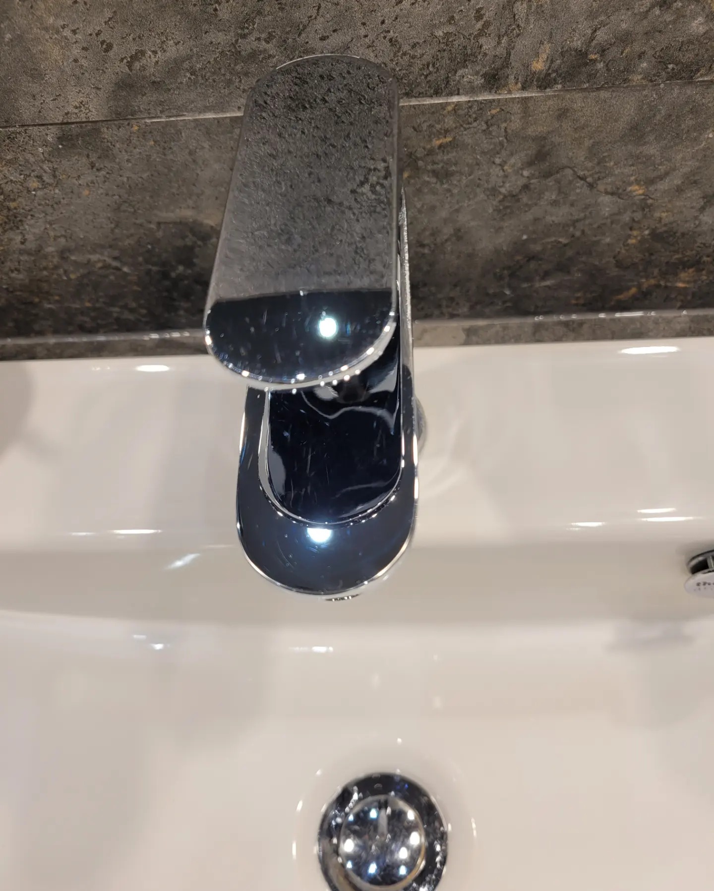 Błyszcząca, chromowana bateria umywalkowa w zbliżeniu, zamontowana na białej umywalce z widocznym odpływem i szarą, kamienną ścianą w tle.