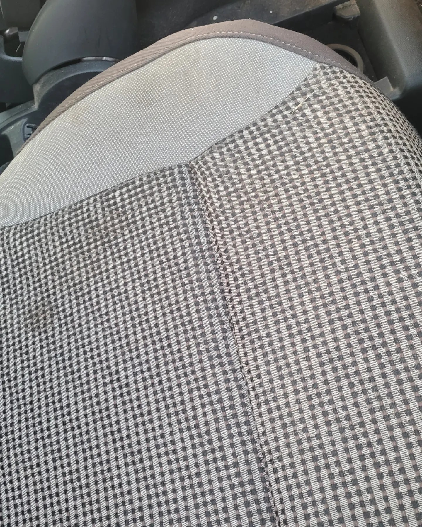 Szczegół fotela samochodowego z szaro-białą tapicerką w drobną kratkę. Widoczny fragment zagłówka i siedziska. Na tapicerce pojedyncze, drobne zabrudzenie.