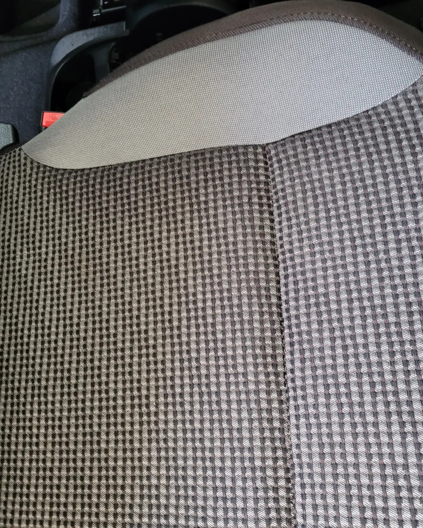 Szczegółowe ujęcie tapicerki fotela samochodowego w kratkę, z widocznym zagłówkiem i fragmentem pasów bezpieczeństwa. Tekstura materiału jest wyraźnie widoczna.