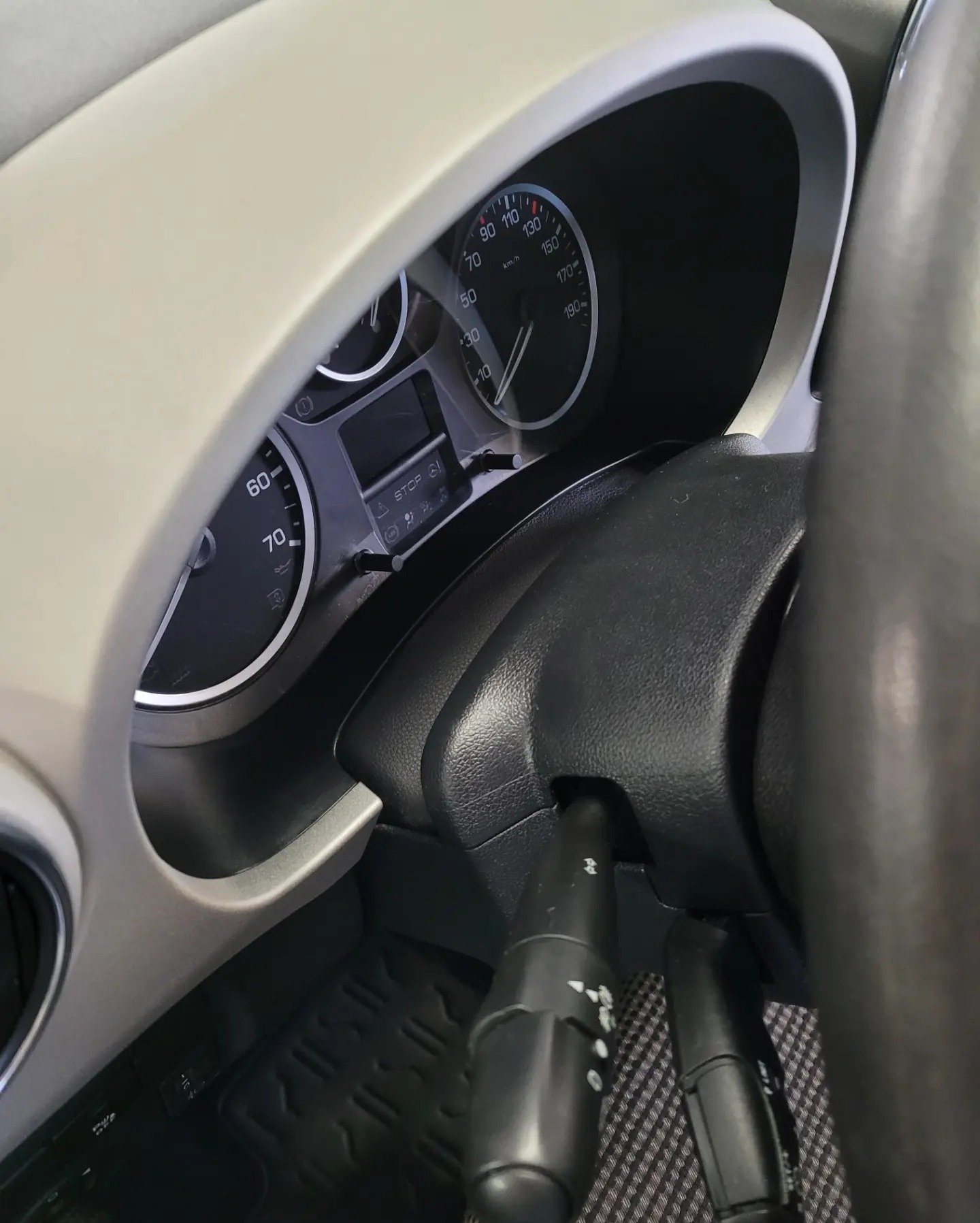Wnętrze samochodu: deska rozdzielcza z prędkościomierzem, kierownica i przełączniki. Widoczna faktura dywaników podłogowych i beżowe wykończenie deski.