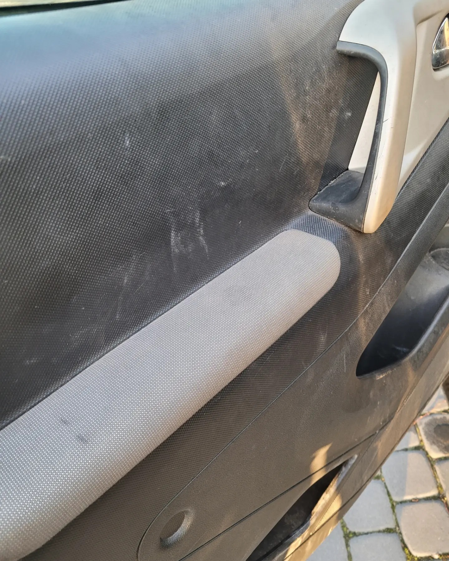 Brudny panel drzwi samochodu z szarą tapicerką i uchwytem. Widoczne ślady użytkowania i zabrudzenia na czarnej, teksturowanej powierzchni.