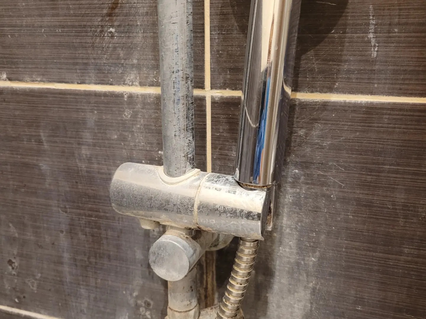 Zabrudzona armatura prysznicowa na tle ciemnych płytek z widocznymi śladami osadów i zacieków. Metalowe elementy pokryte warstwą brudu i kamienia.